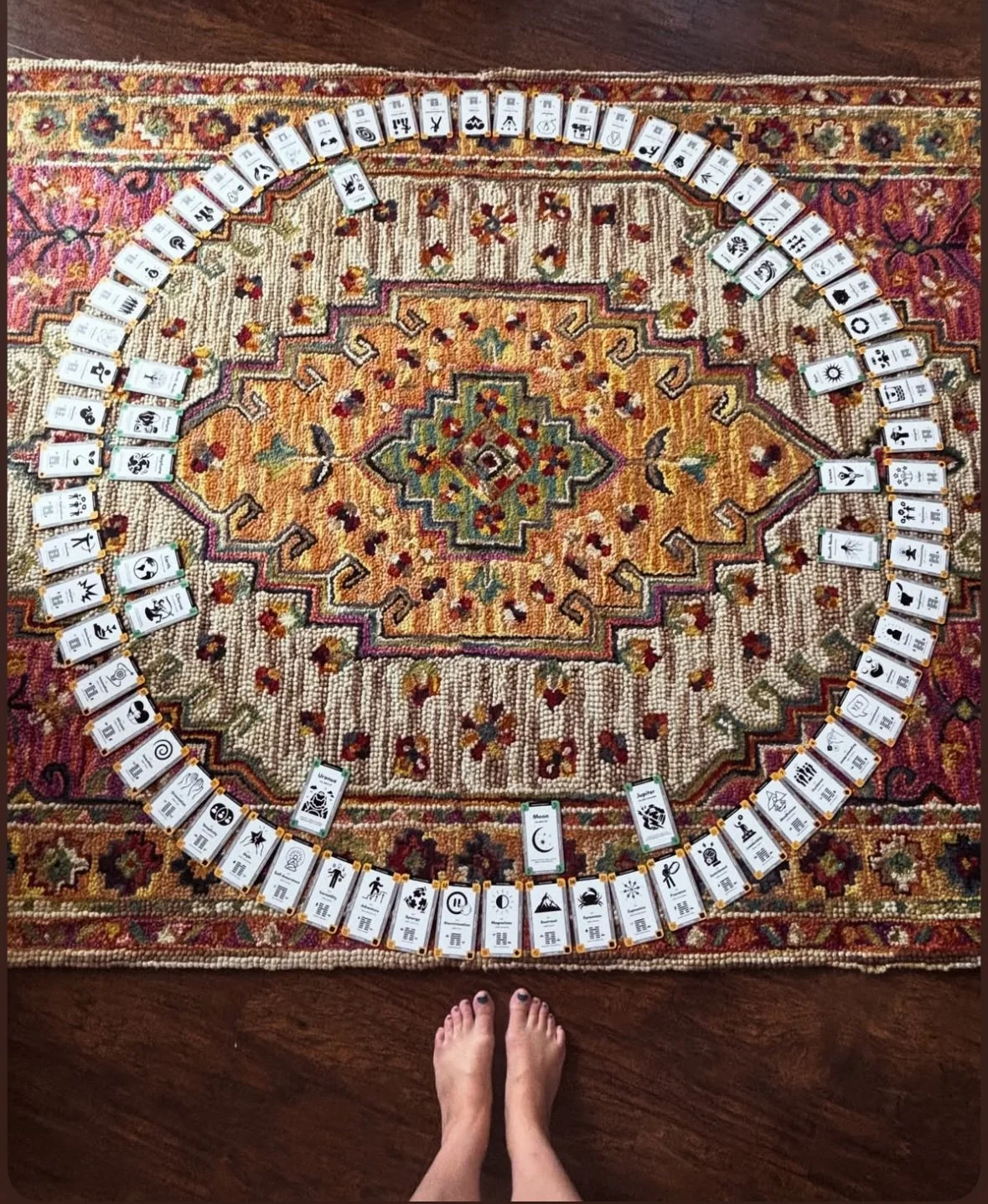 A person with painted toenails standing on a patterned area rug, with cards laid out in a circle on the rug.