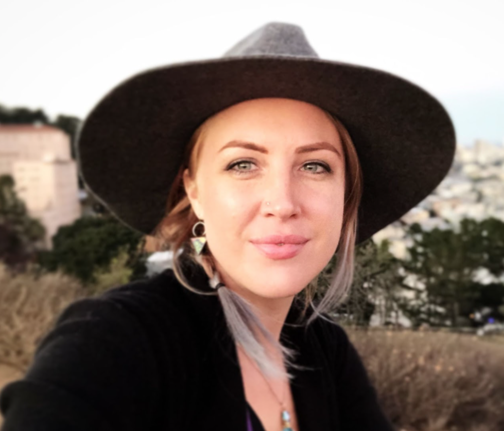 A woman with fair skin and blue eyes wearing a large brown hat and a black top, standing outdoors with trees and city buildings in the background.