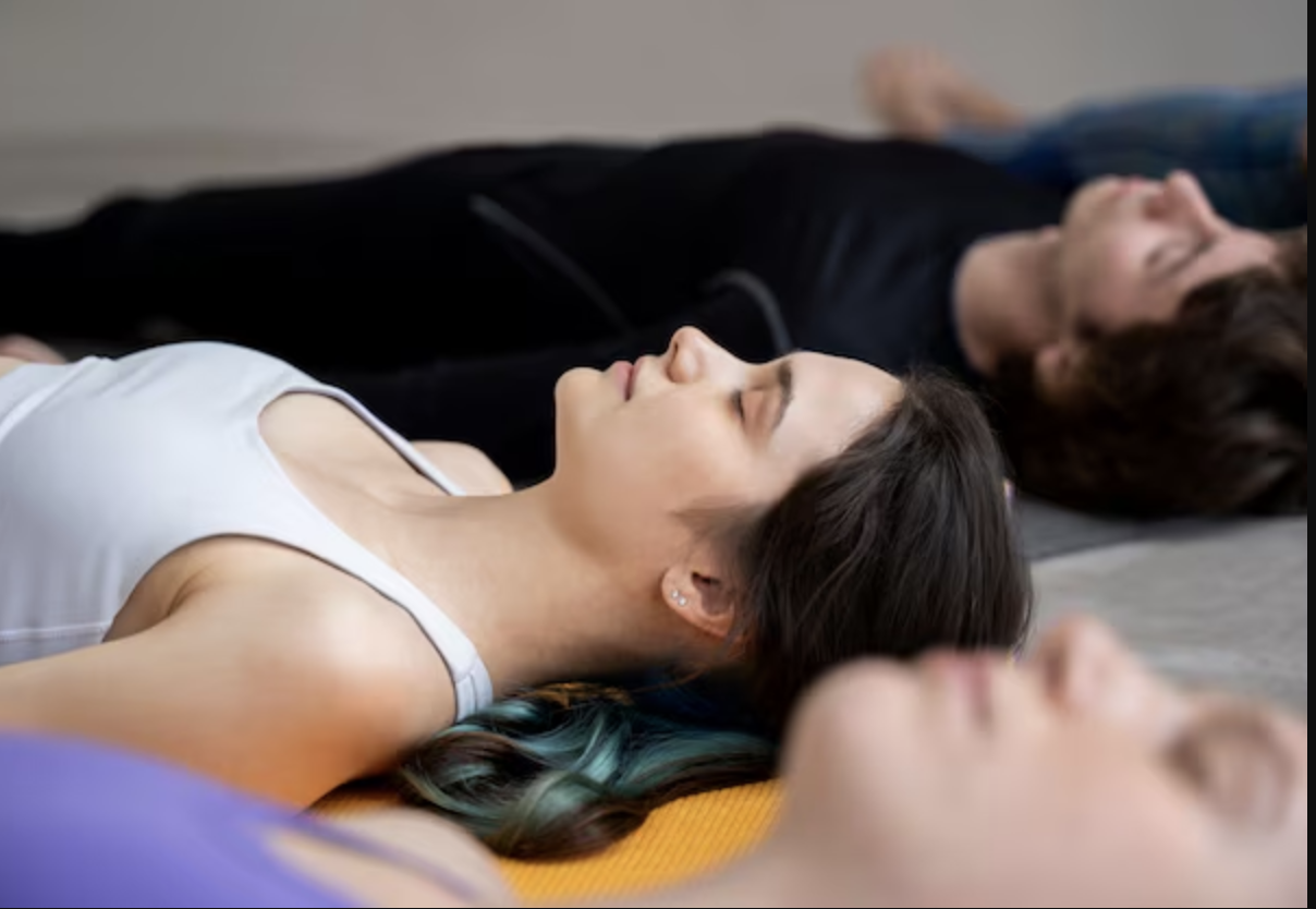 People lying on yoga mats, eyes closed, in a meditation or relaxation pose.