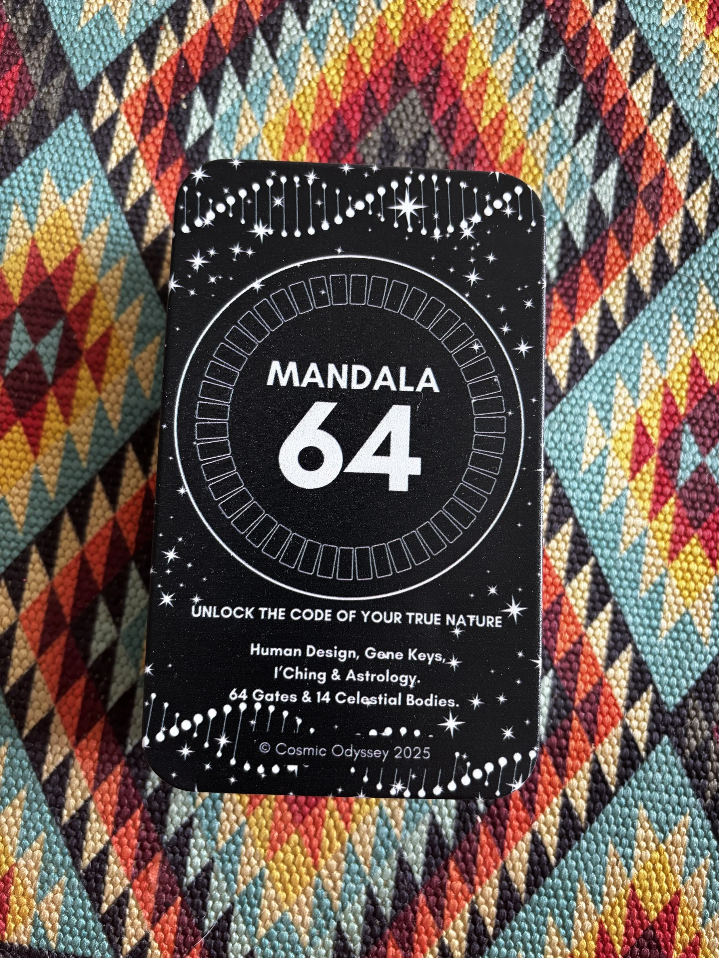 Black card titled "Mandala 64" with white star-like designs, celestial motifs, and text about unlocking human design, gene keys, astrology, and celestial bodies. The card is placed on a colorful woven fabric with geometric patterns.