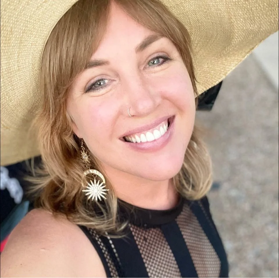 A woman with shoulder-length blonde hair is smiling and wearing a wide-brimmed straw hat and large, starburst-shaped earrings.