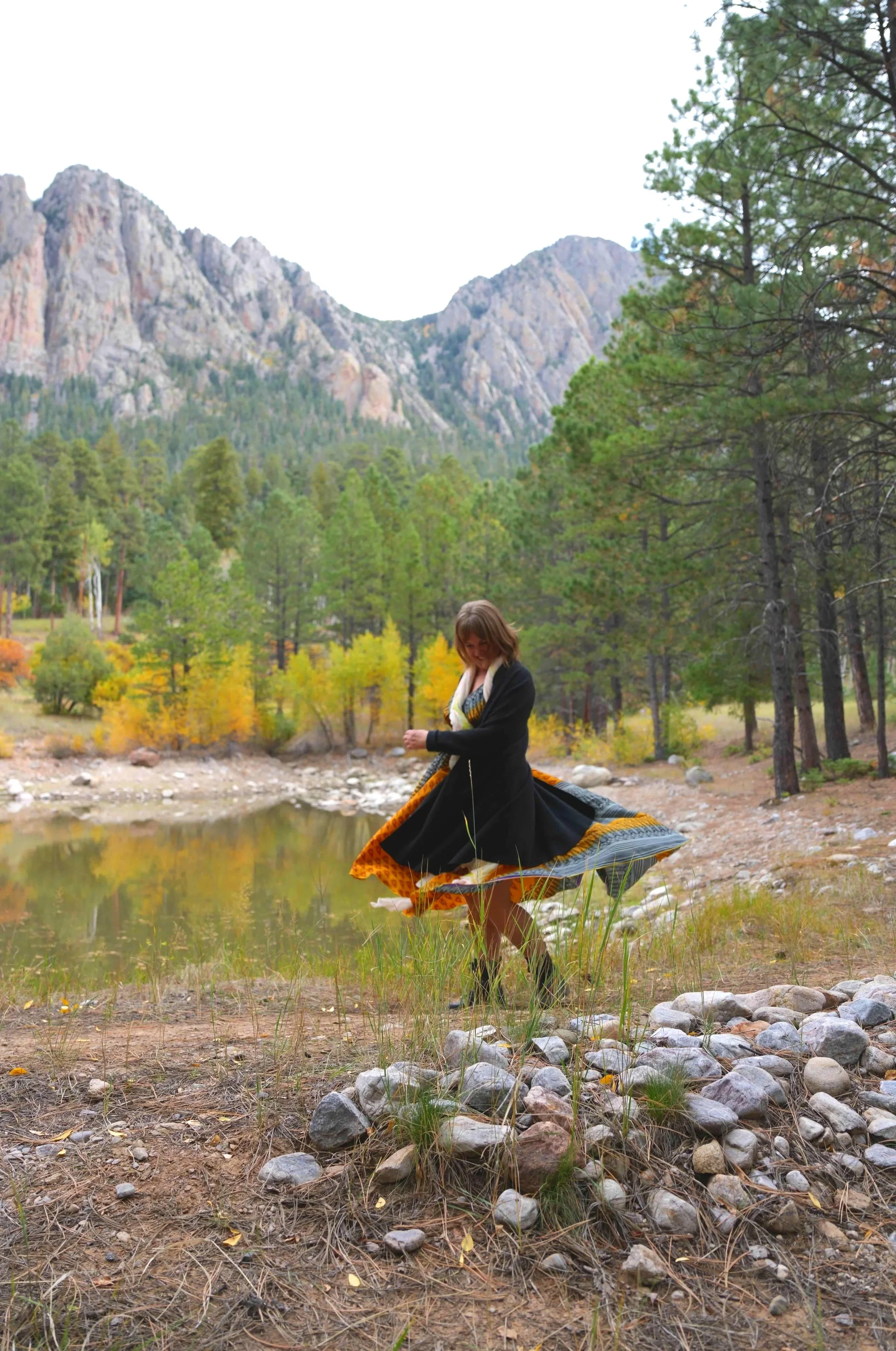 A woman in a black dress with an orange and black underskirt spinning near a lake in a forested area with mountains in the background.
