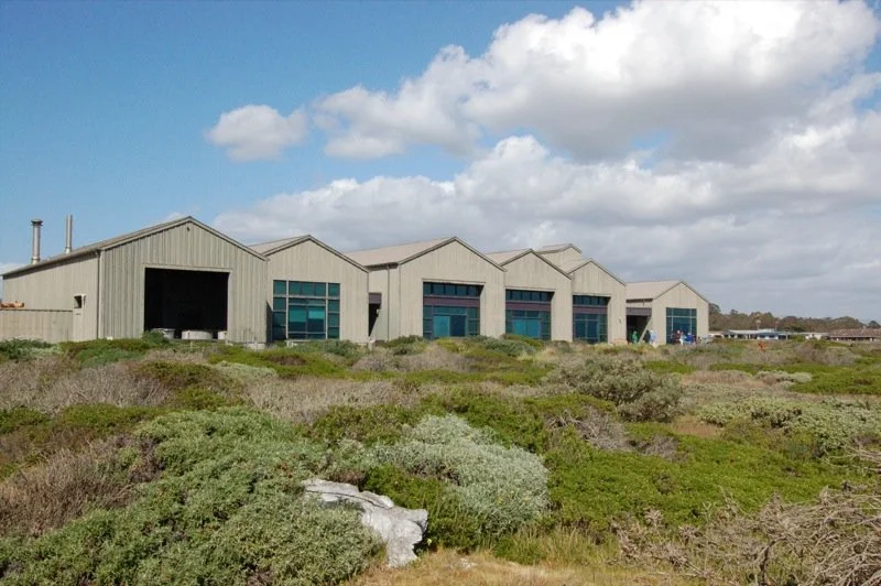 Large industrial warehouse building with multiple garage doors surrounded by native bushes and shrubs, under a cloudy sky.