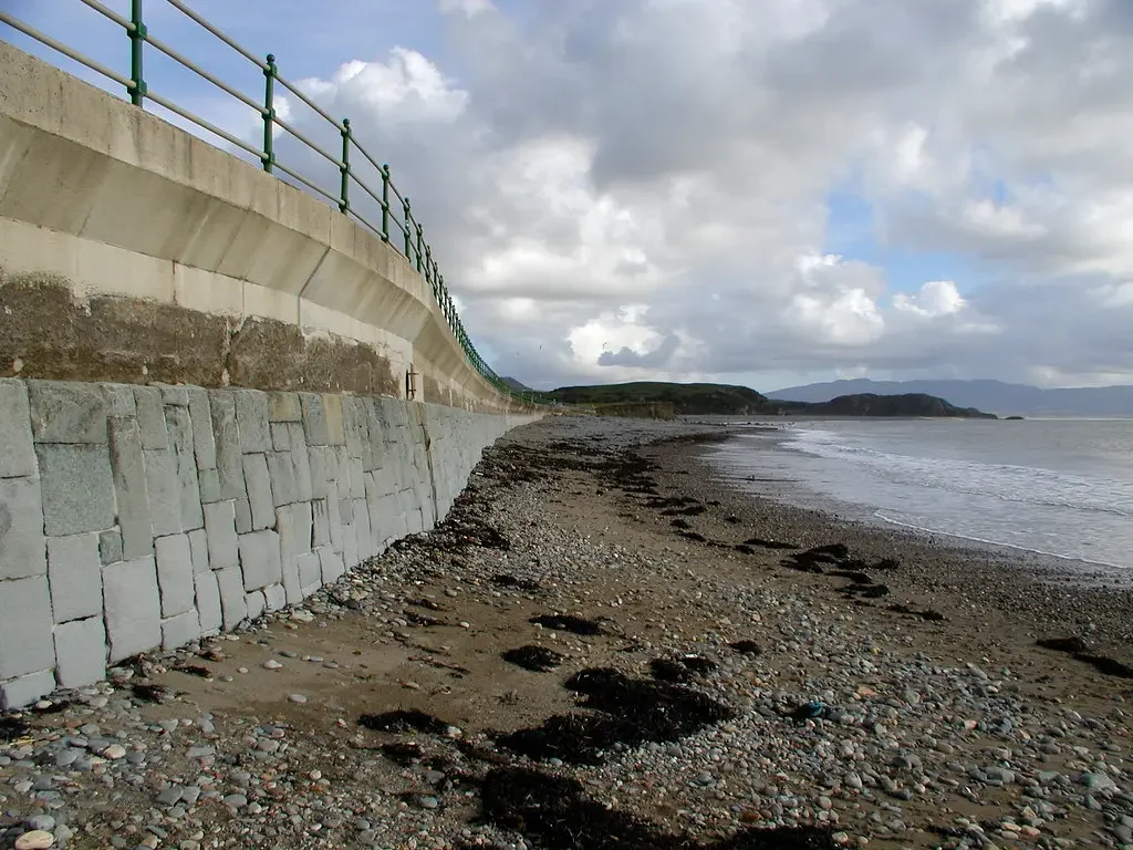 View of a beach with a stone and sand shoreline, seaweed on the sand, a concrete sea wall with green railings, and hills in the background under a cloudy sky.