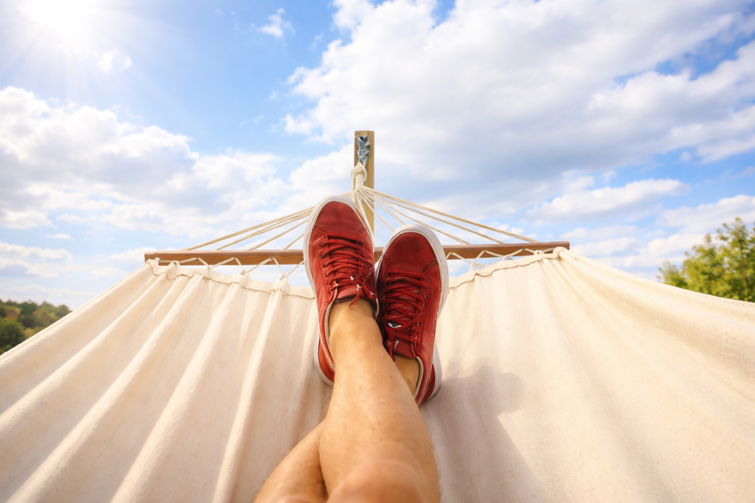 Person wearing red sneakers relaxing on a hammock outdoors, with a partly cloudy sky above.