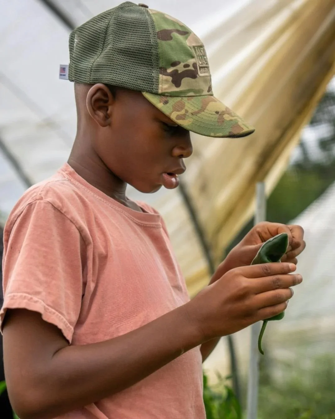 A profile view of a young student wearing a camouflage baseball cap, carefully holding and observing a large green leaf in an outdoor learning environment.
