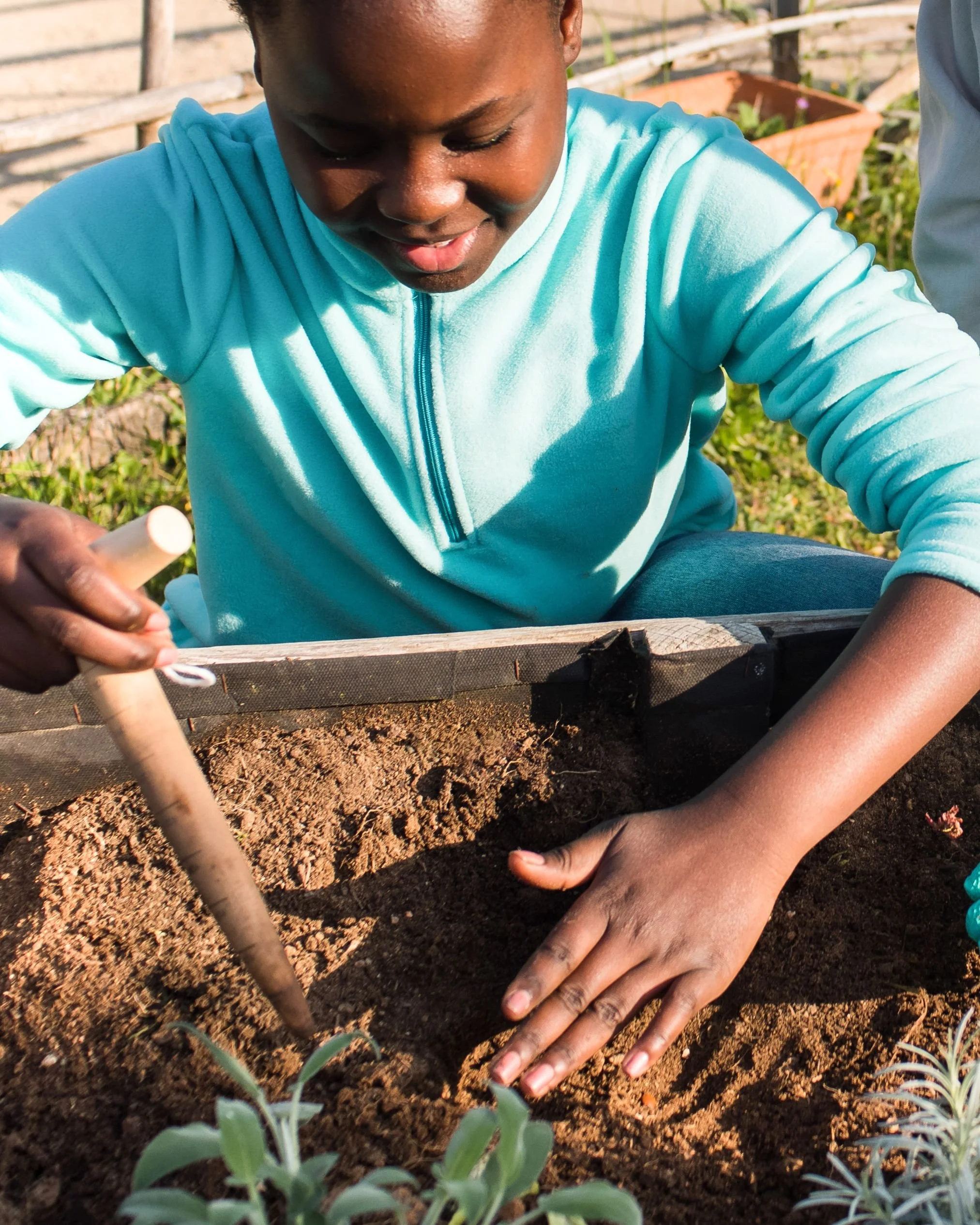 A young student in a turquoise fleece smiling while using a small hand tool to prepare soil in a wooden raised garden bed for planting.