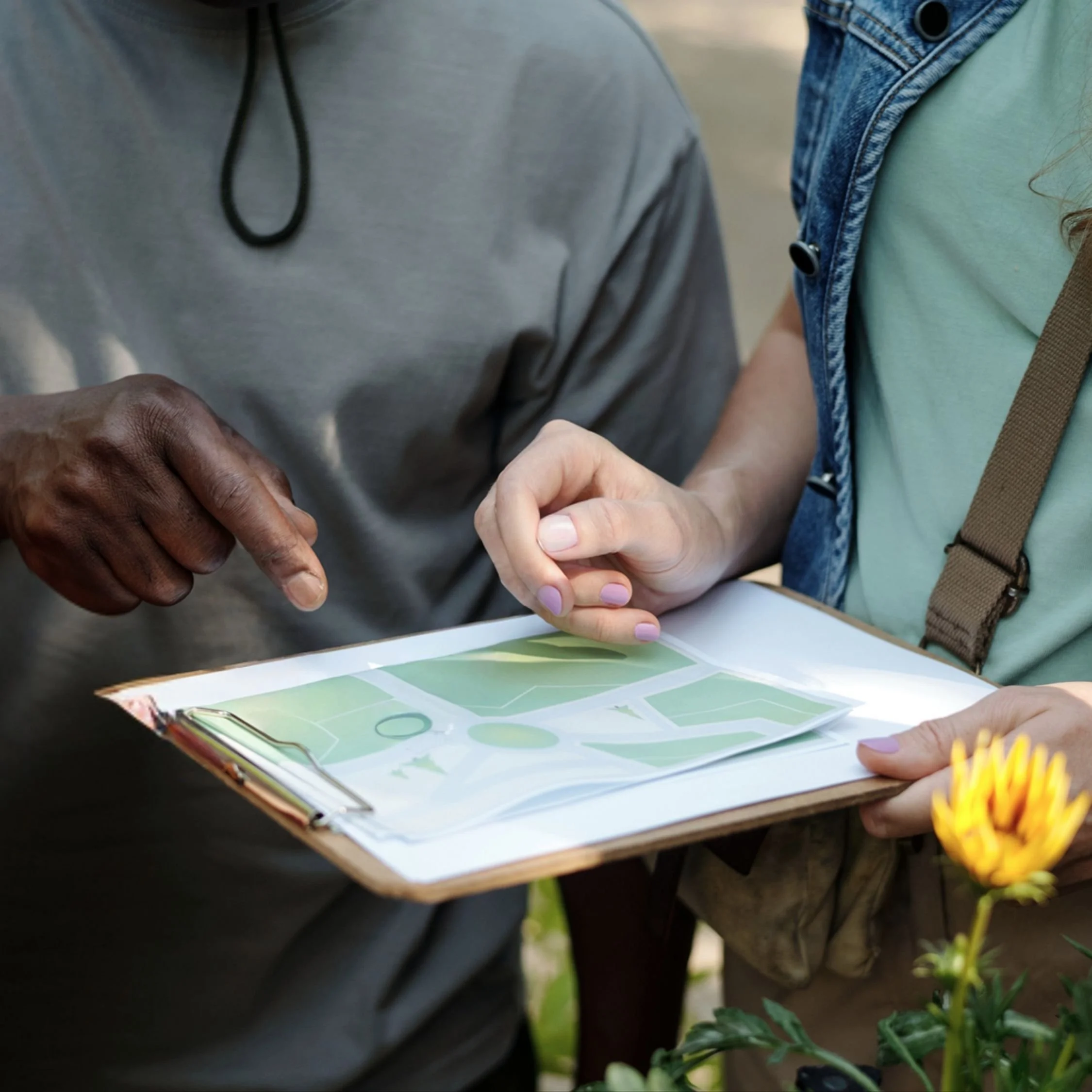 Close-up of two individuals reviewing a colorful landscape architectural plan on a clipboard while standing outdoors near green foliage.