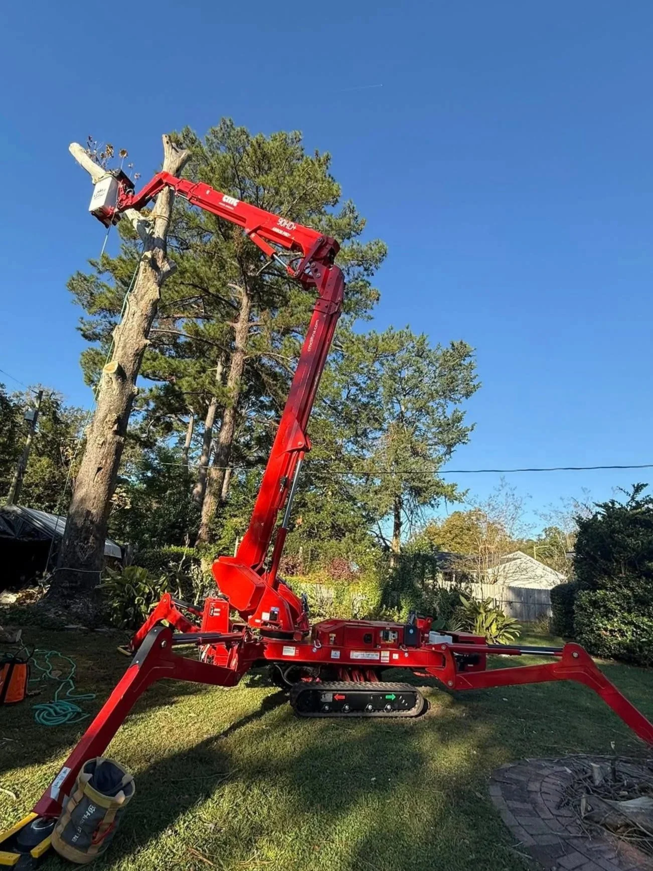 A red bucket truck with a long extending arm is trimming trees in a residential yard under a clear blue sky.