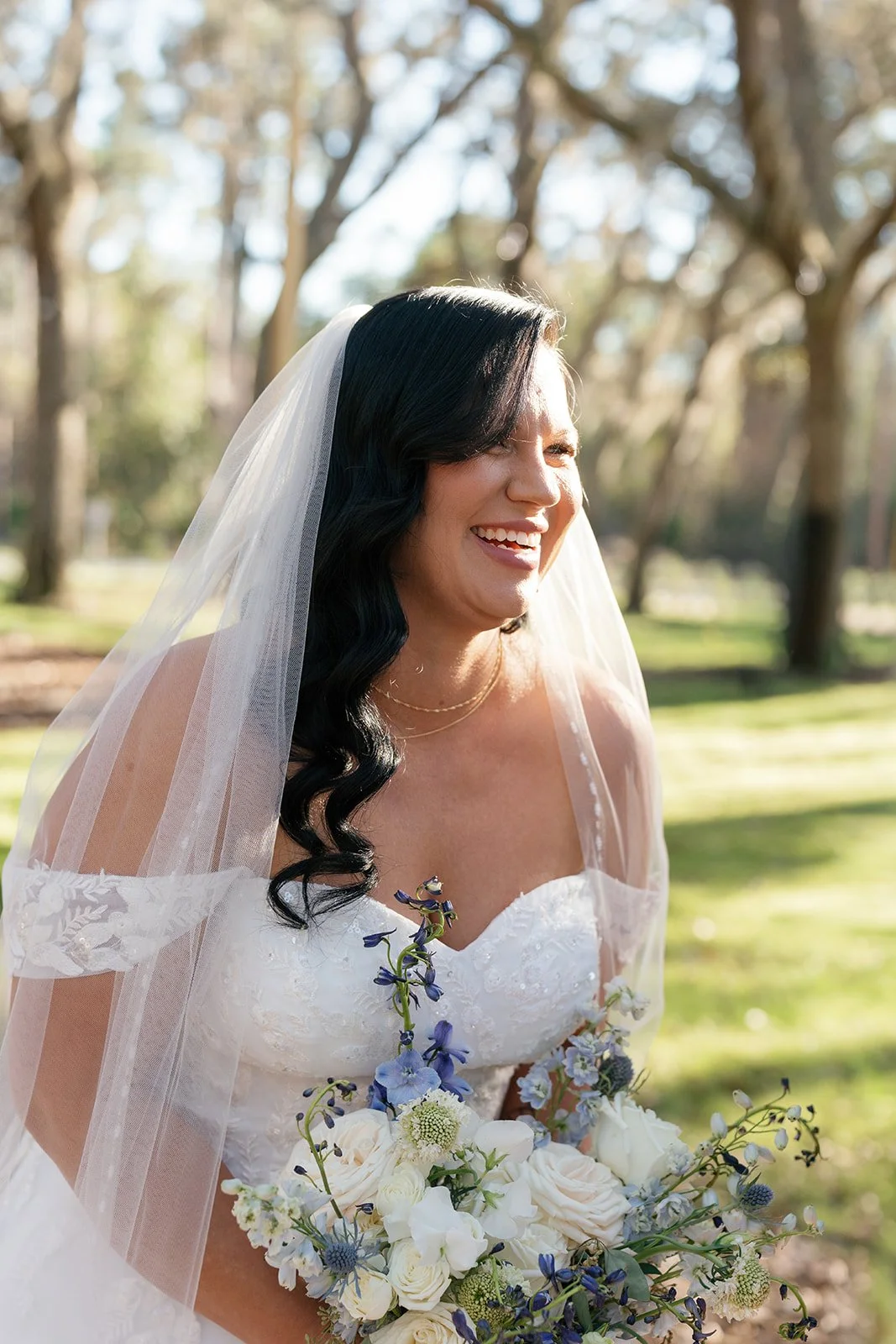 A bride in a white wedding dress with a veil, smiling and holding a bouquet of white and purple flowers, standing outdoors in a park with trees.