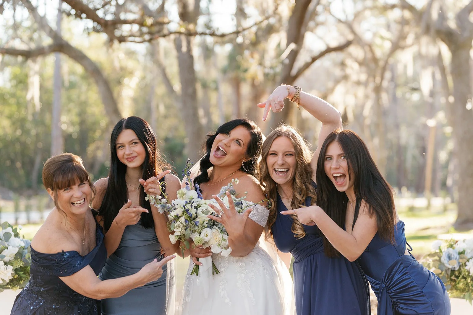 Group of five women celebrating at outdoor wedding, with trees and sunlight in the background, one holding a bouquet, others pointing and smiling.
