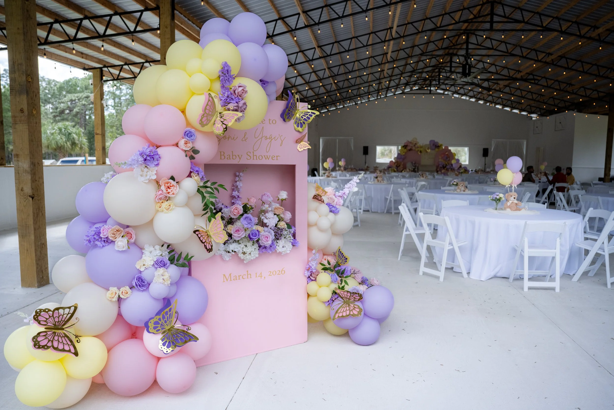 Decorative balloon arch in pastel colors with purple and yellow balloons, flower arrangements, and butterfly decorations at a baby shower event.