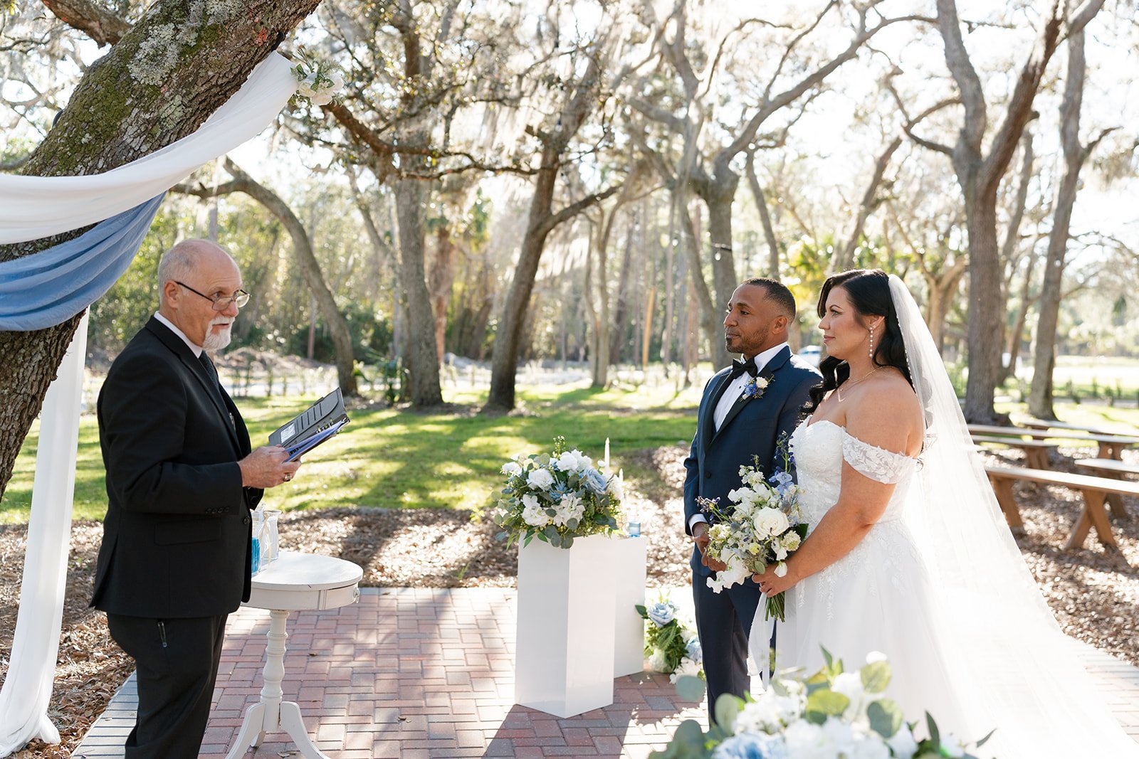 A bride and groom standing at an outdoor wedding ceremony, facing an officiant who is reading from a book. The bride wears a white off-the-shoulder wedding gown with a veil, and the groom wears a navy blue tuxedo with a bow tie. They hold bouquets of