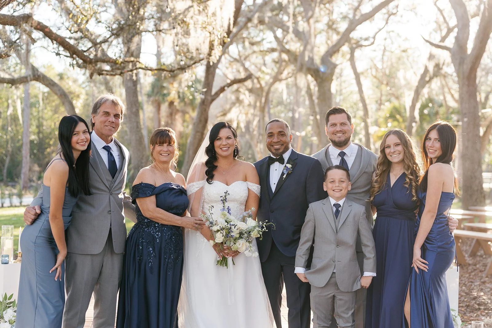 Group of nine people dressed in formal wedding attire, standing outdoors in a wooded area with sunlight filtering through the trees, celebrating a wedding.