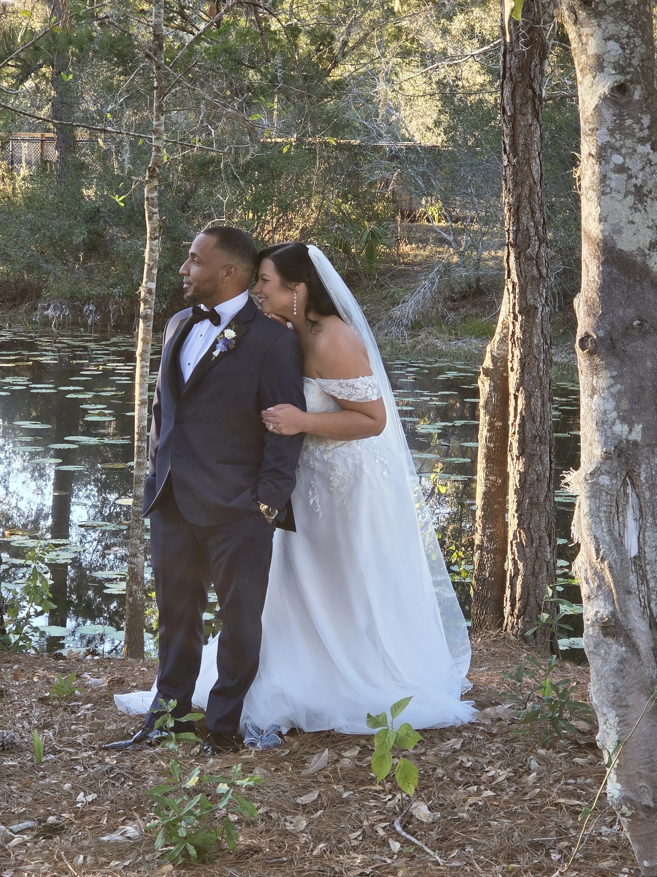 A newlywed couple standing by a pond in a forested area. The groom in a black tuxedo and the bride in a white lace wedding dress and veil. Trees and water lilies are visible in the background.
