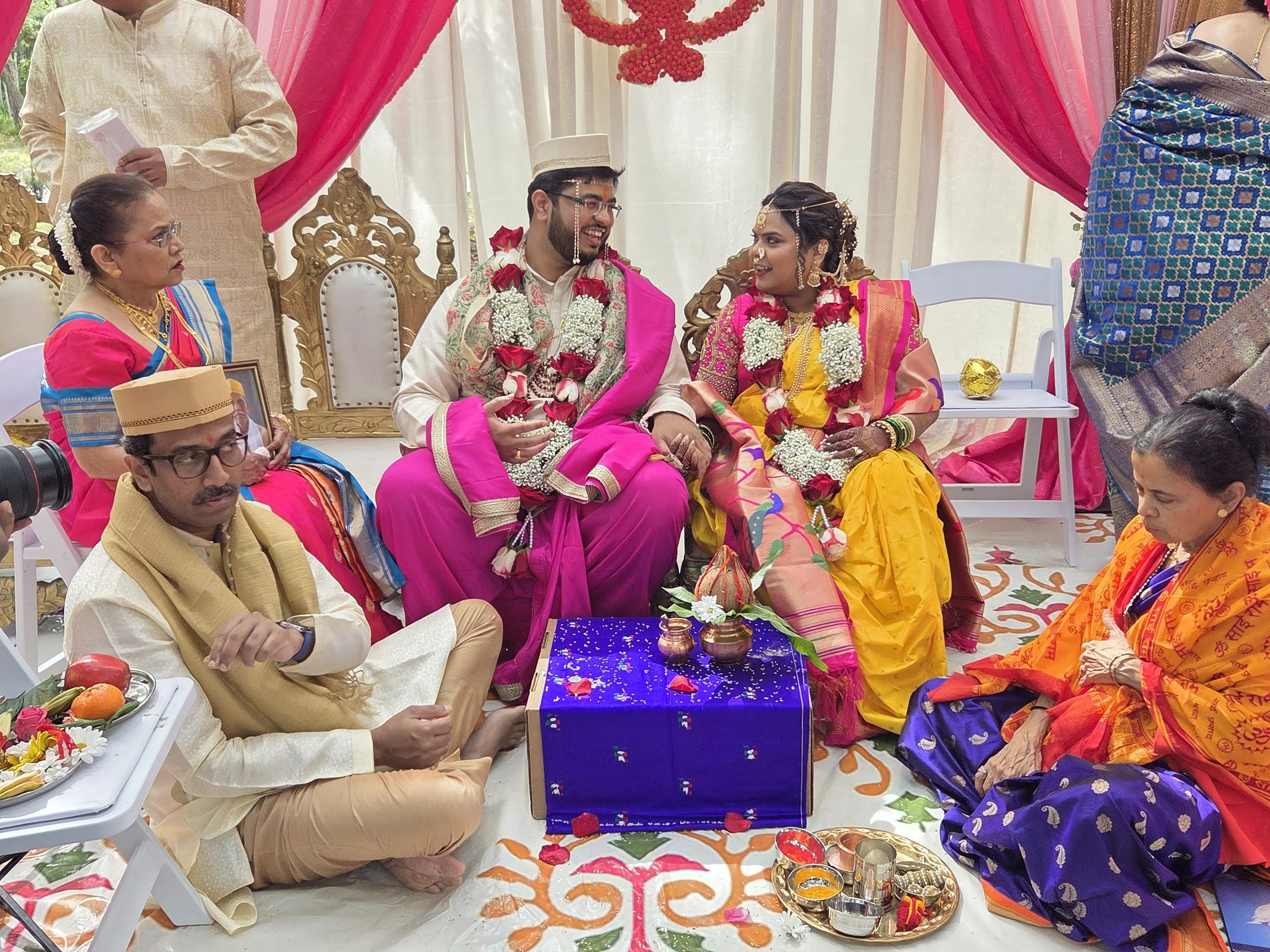 Hindu wedding ceremony with the bride and groom seated on ornately carved wooden chairs, surrounded by family members in traditional colorful attire under a decorated canopy.
