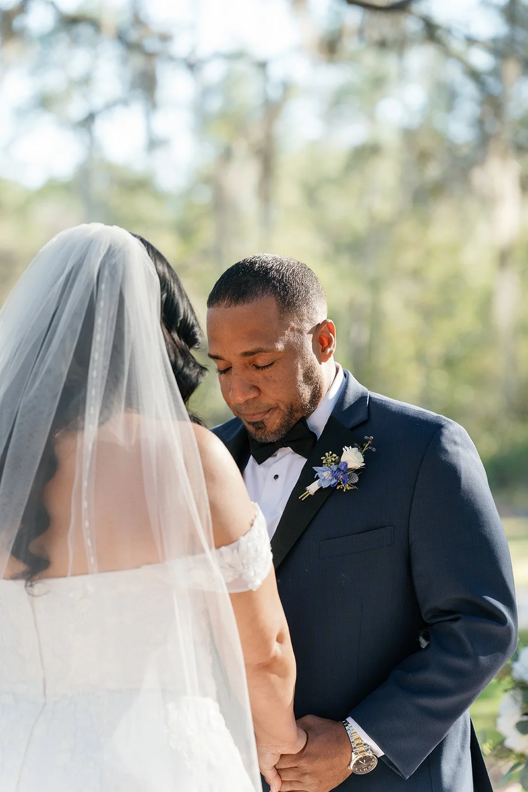 A couple getting married outdoors, with the bride wearing a veil and off-shoulder dress, and the groom in a tuxedo with a boutonnière, holding hands and with eyes closed.