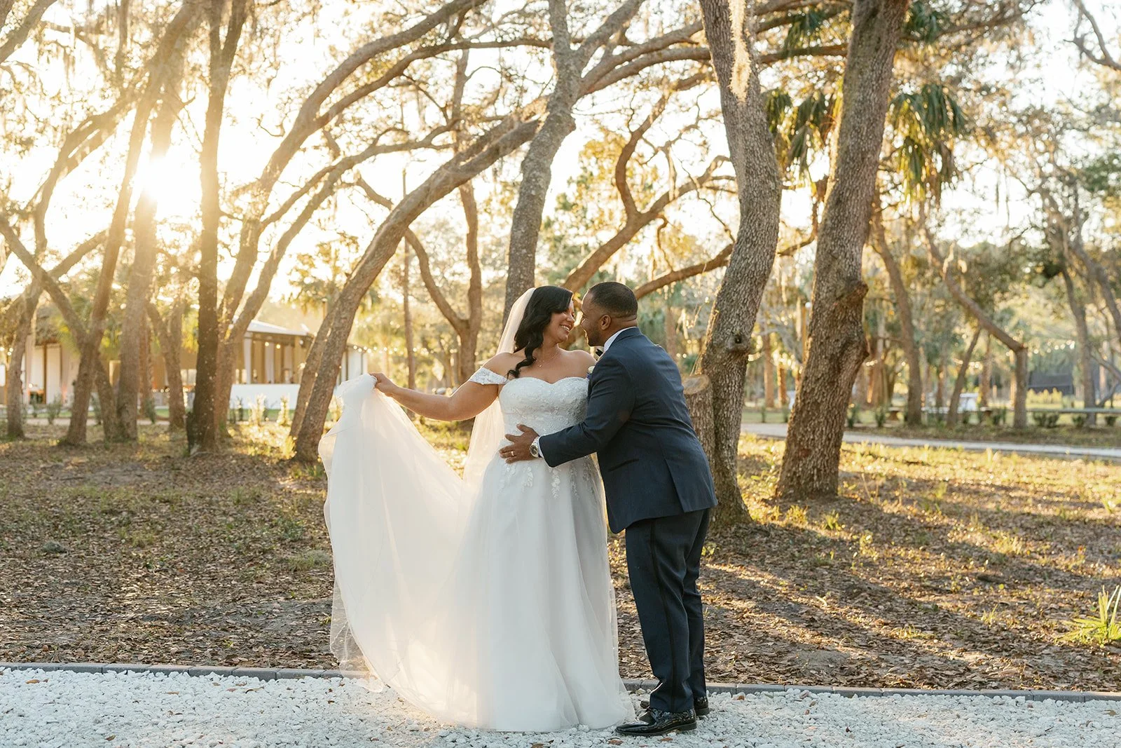 A bride and groom holding hands and smiling at each other in a park with trees and sunlight.