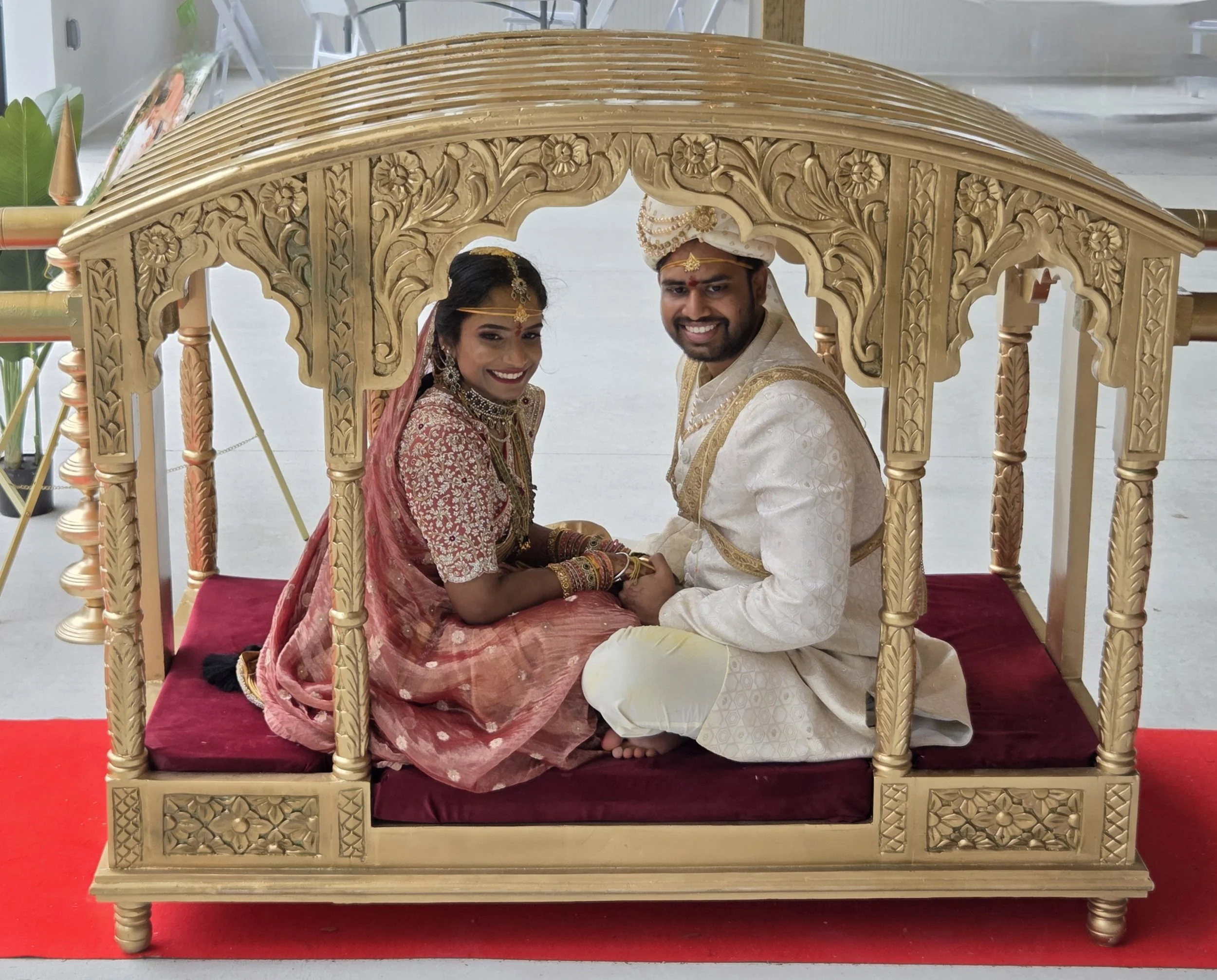 A bride and groom dressed in traditional Indian wedding attire, sitting on a wedding palanquin with intricate carvings, inside a building with a red carpet underneath.