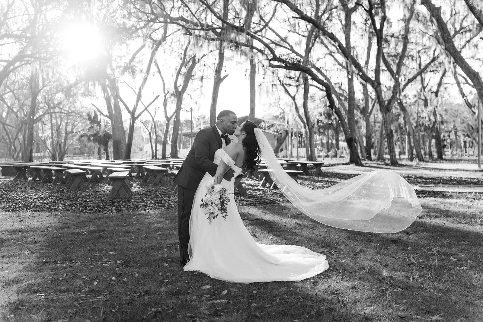 A bride and groom sharing a kiss outdoors, with the bride holding a bouquet and wearing a long veil, in a park with trees and benches in the background.