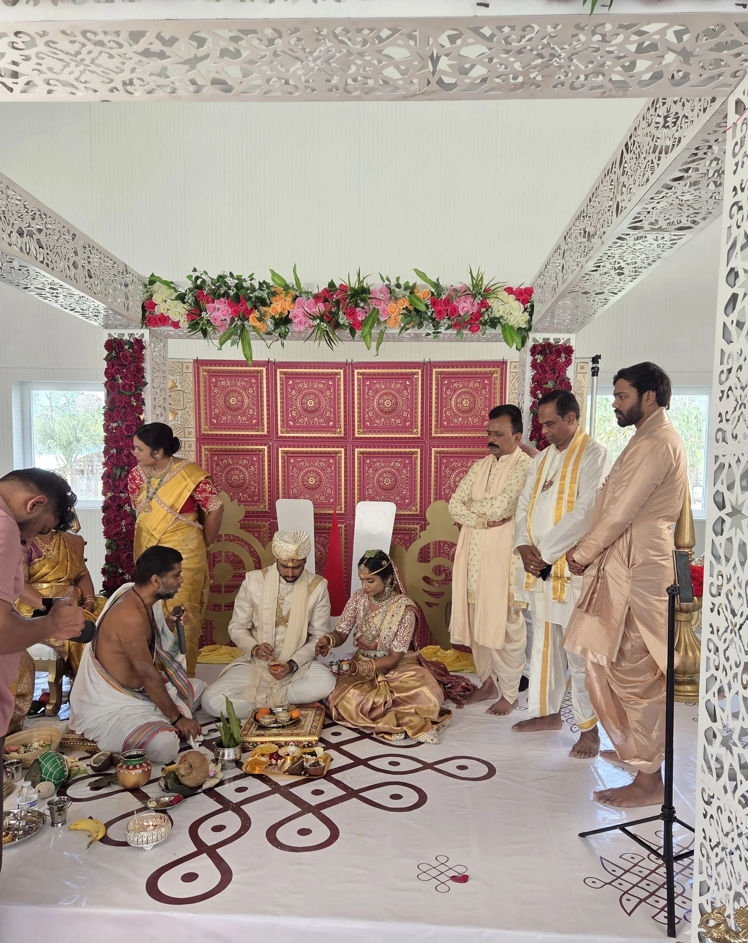 A traditional Hindu wedding ceremony with a bride and groom seated on the floor, surrounded by family members and priests. The altar is decorated with flowers, intricate woodwork, and artwork. Ritual items are placed on the floor in front of the coup