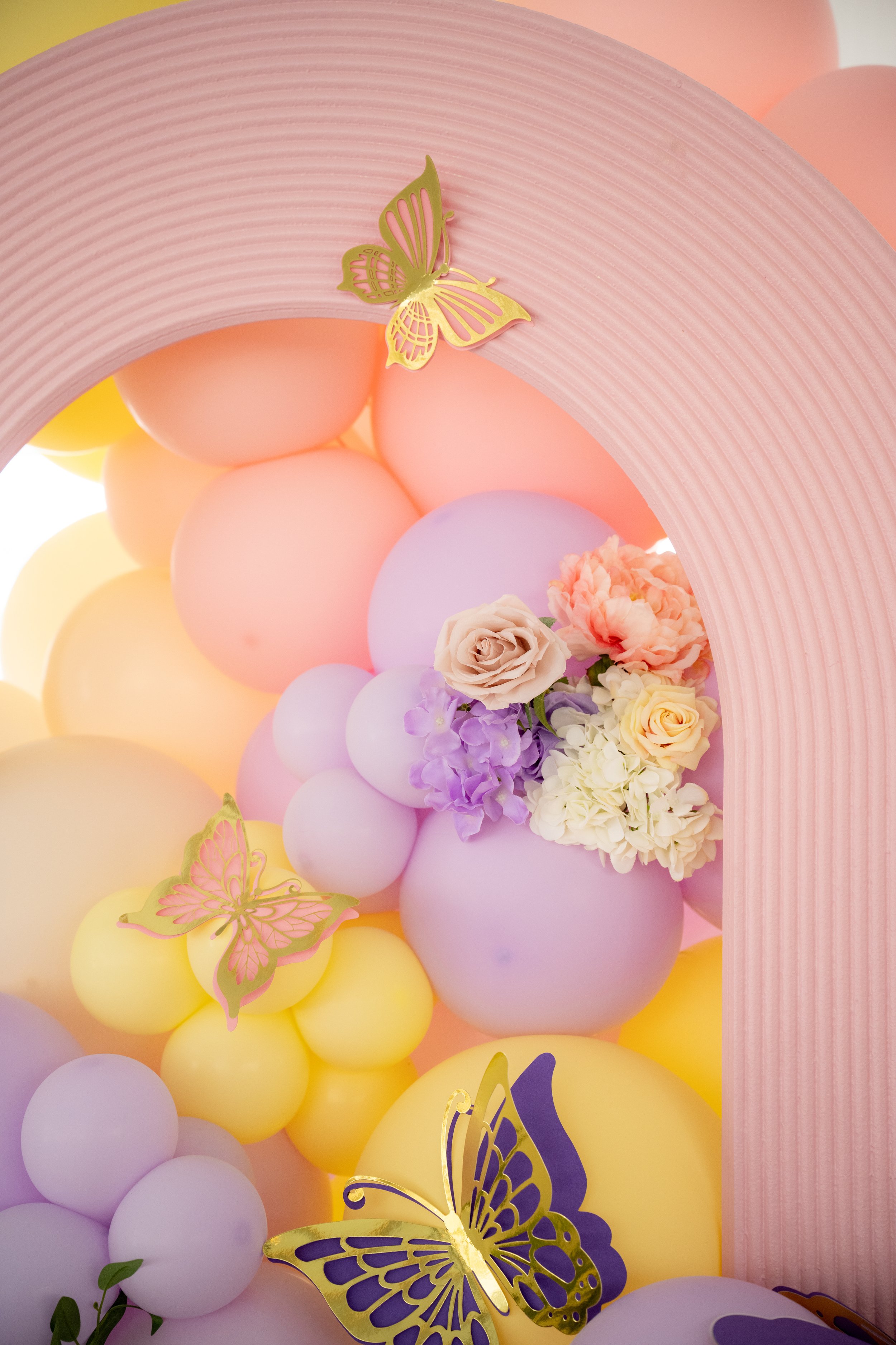 Decorative balloon arch with pastel-colored balloons, pink paper backdrop, and gold and purple butterfly accents, with floral arrangements at the base.