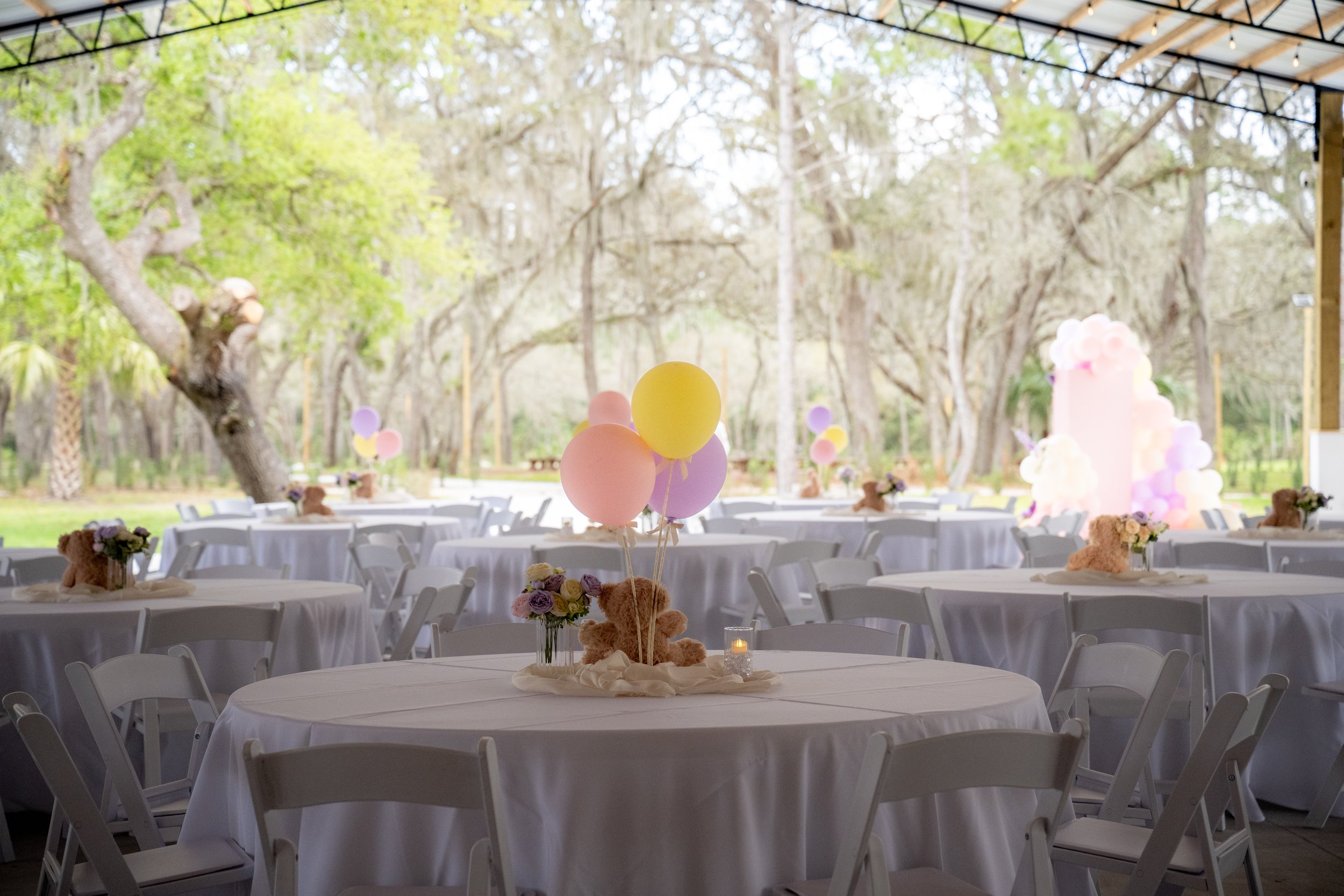 Outdoor event space decorated with pastel balloons, teddy bears, and flower arrangements on tables, with trees and a metal canopy overhead.