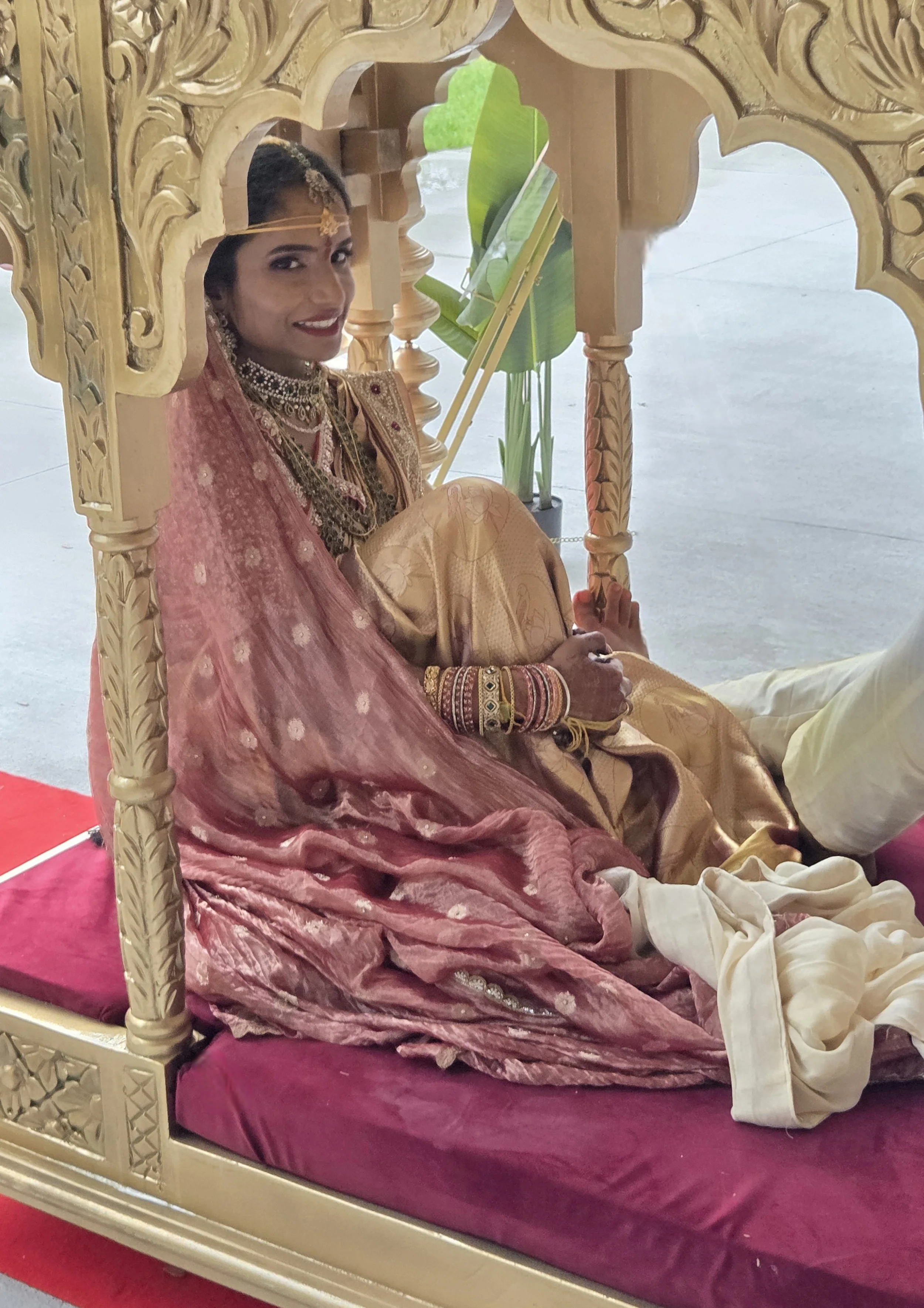 A woman dressed in traditional Indian bridal attire sitting on a decorated mattress under a golden canopy with intricate carvings. She is wearing jewelry and a pink saree, smiling, with green leaves in the background.