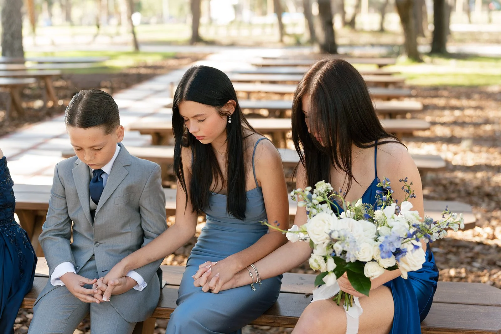 Three people sit on a park bench with their heads bowed and eyes closed, appearing to pray or reflect. The first person is a young boy in a gray suit, the second is a woman in a blue dress, and the third is a woman in a darker blue dress holding a bo