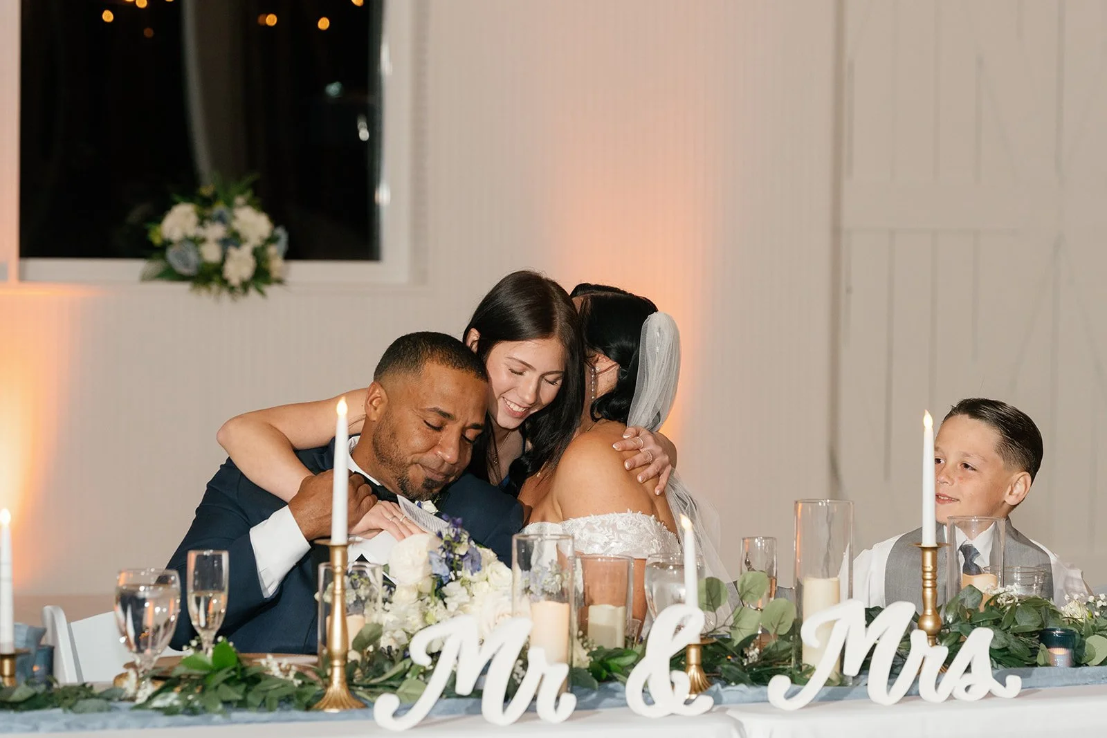 A wedding reception scene with four people, two men and two women. The bride and groom are hugging, and the bride is wearing a white dress with an off-the-shoulder neckline. The groom is dressed in a dark suit, and one of the women has black hair and