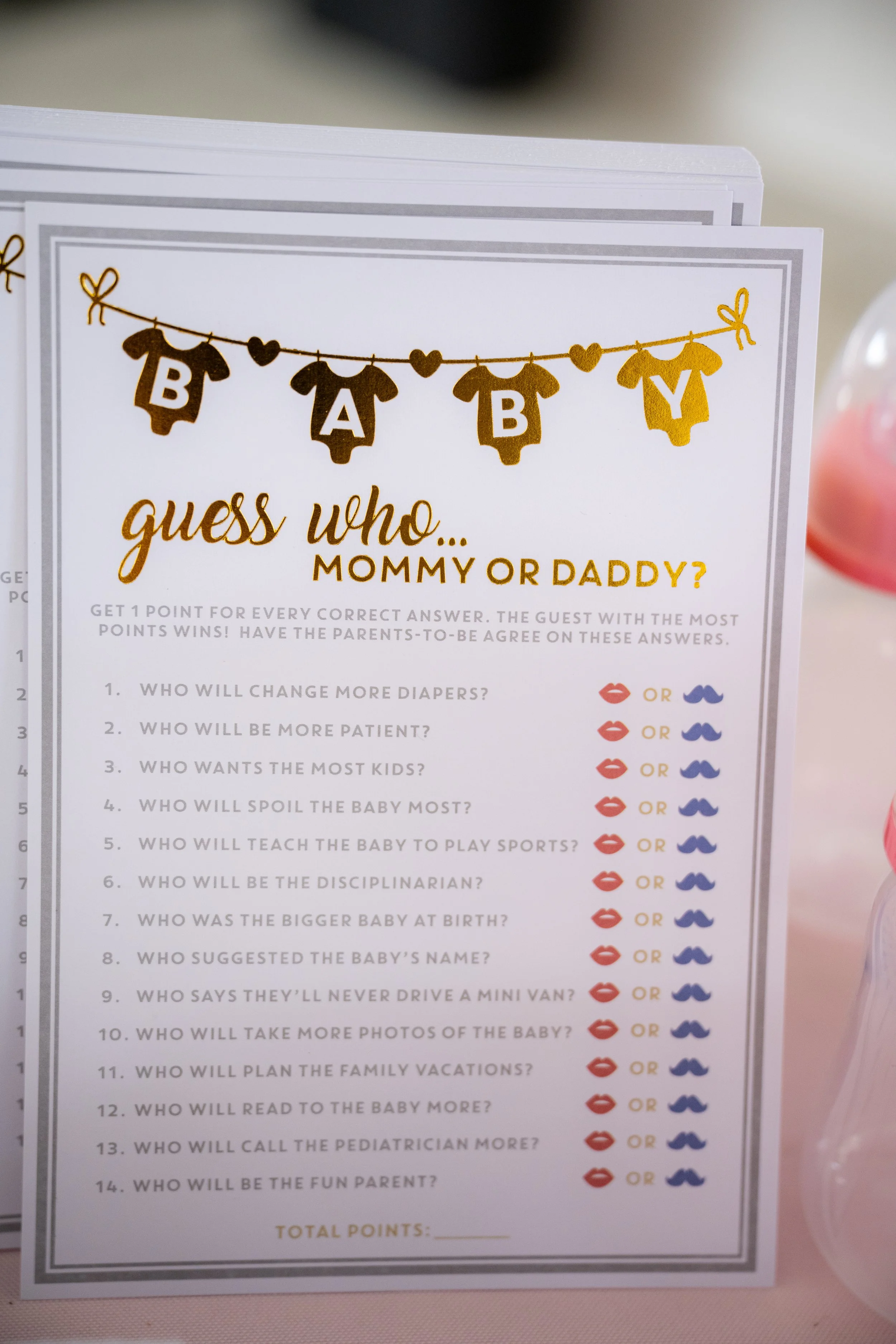 A parenting game card titled 'Guess Who... Mommy or Daddy?' decorated with gold Bodysuit cutouts spelling 'BABY' hanging on a string with heart accents. The card lists questions for a guessing game about parental roles and habits.