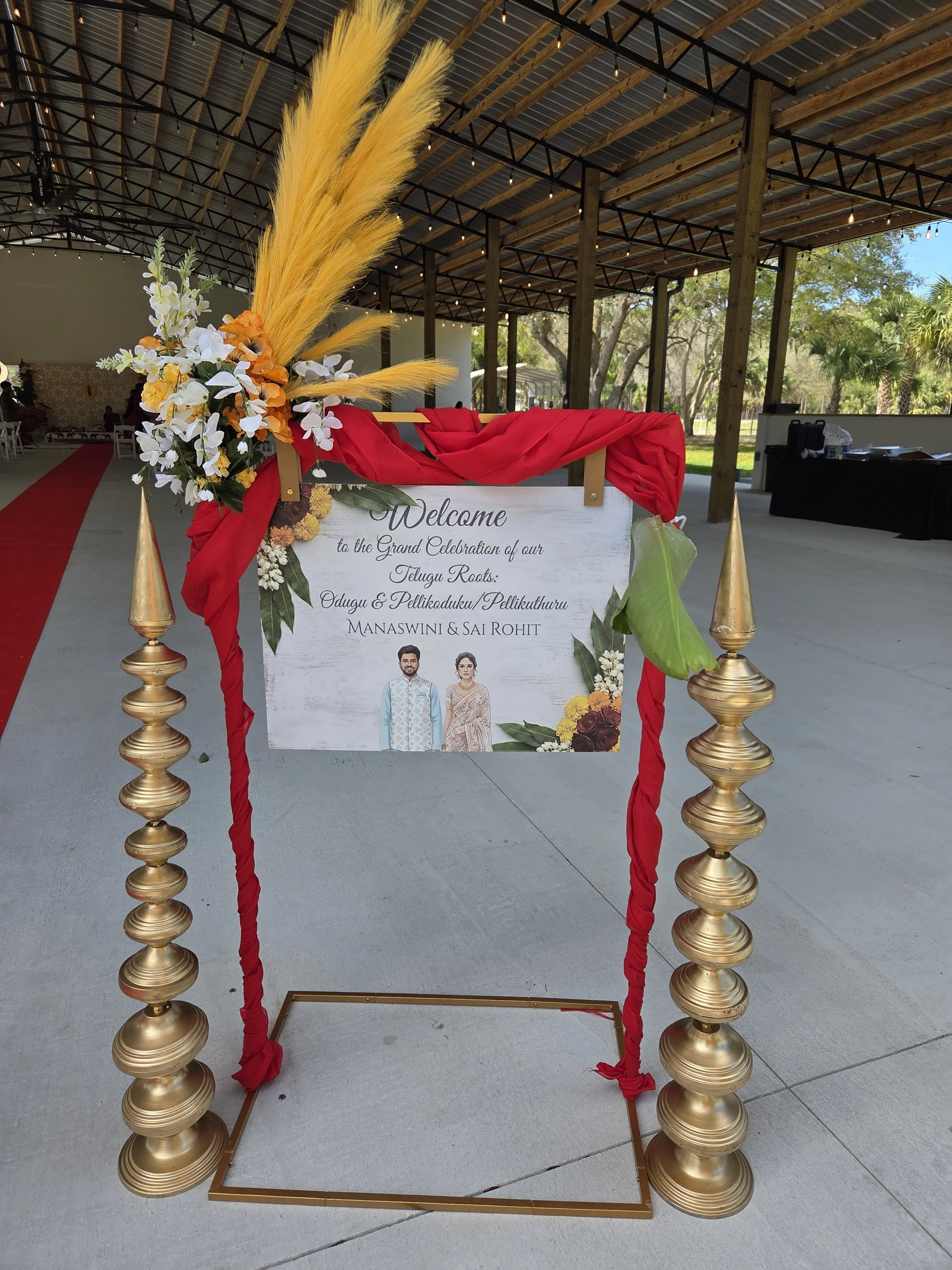 Decorative wedding sign with floral arrangement on a stand, reading 'Welcome to the Grand Celebration of our Telugu Roots, Odugu & Pelikaduku/Pellikuthuru, Manaswini & Sai Rohit,' at an indoor event space with trees visible outside.