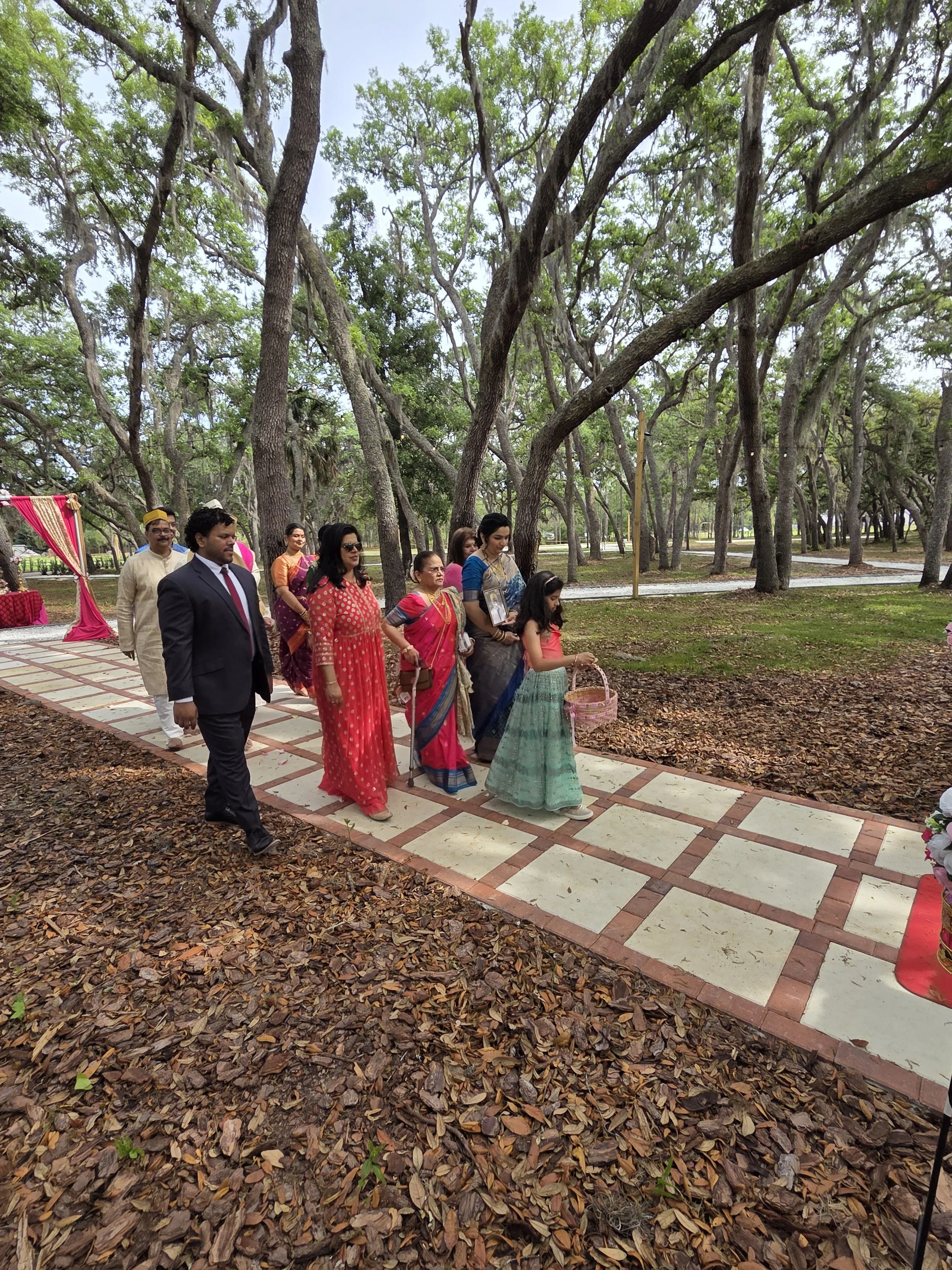 People participating in a wedding ceremony outdoors in a wooded area, walking along a brick pathway covered with fallen leaves, wearing traditional Indian attire.