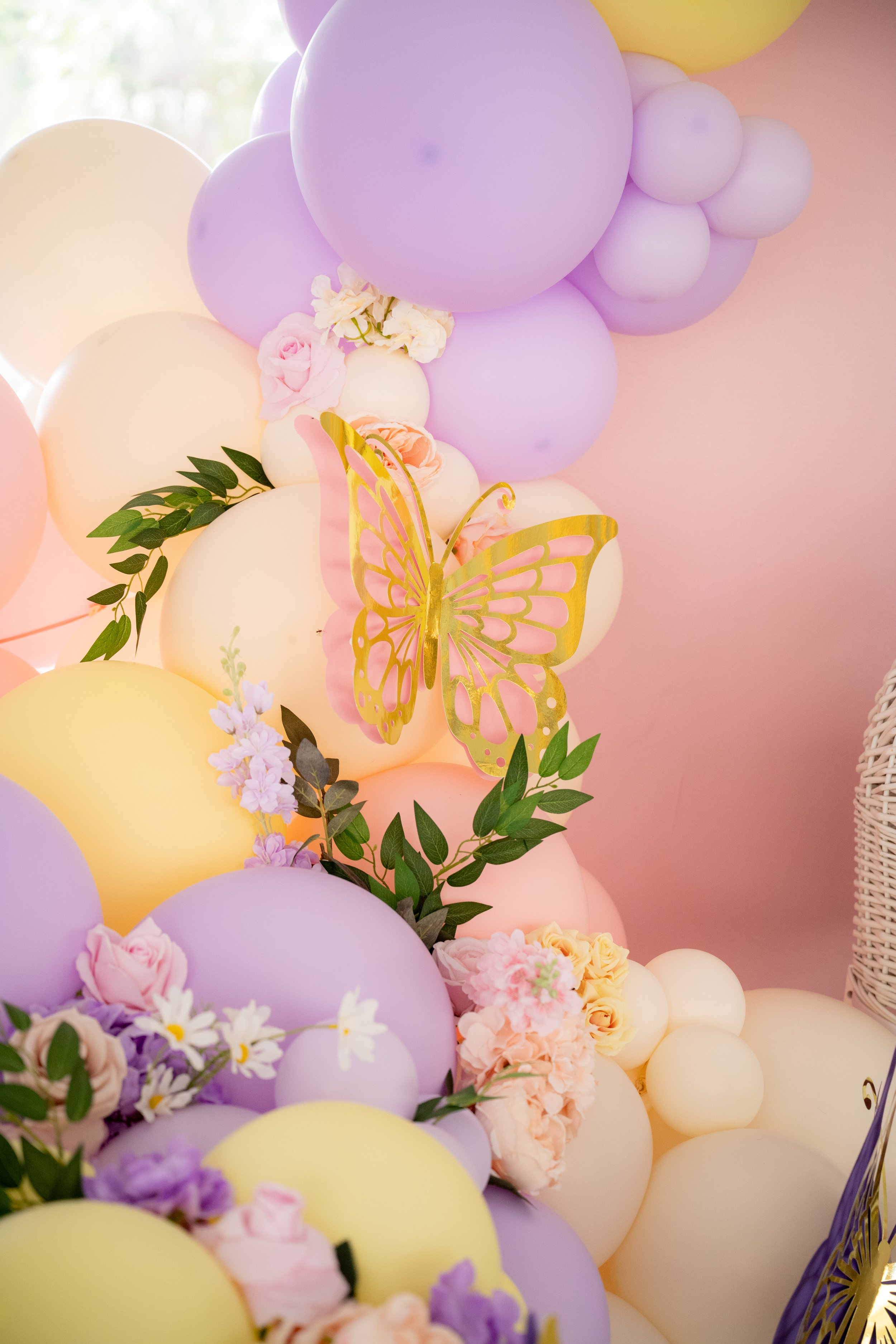 Decorative arrangement with pastel-colored balloons, pink and yellow flowers, greenery, and gold butterfly cutout.