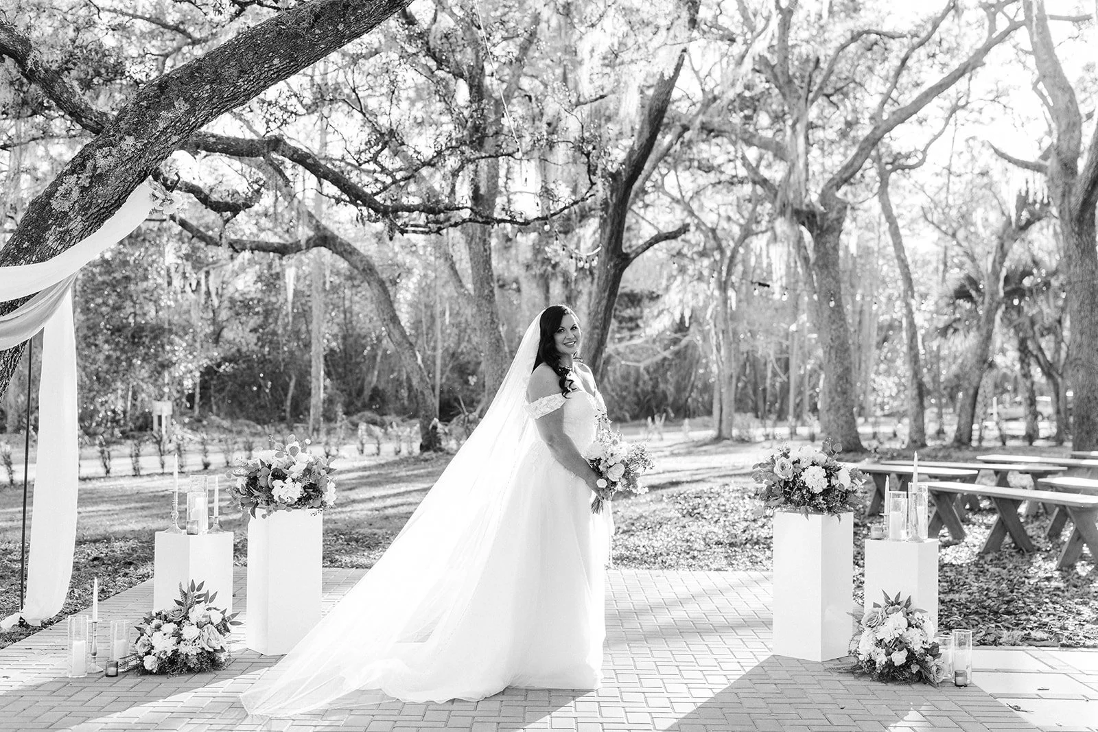 A bride in a wedding dress holding a bouquet, standing outdoors during daytime in a park with trees and benches, decorated with flowers and candles.