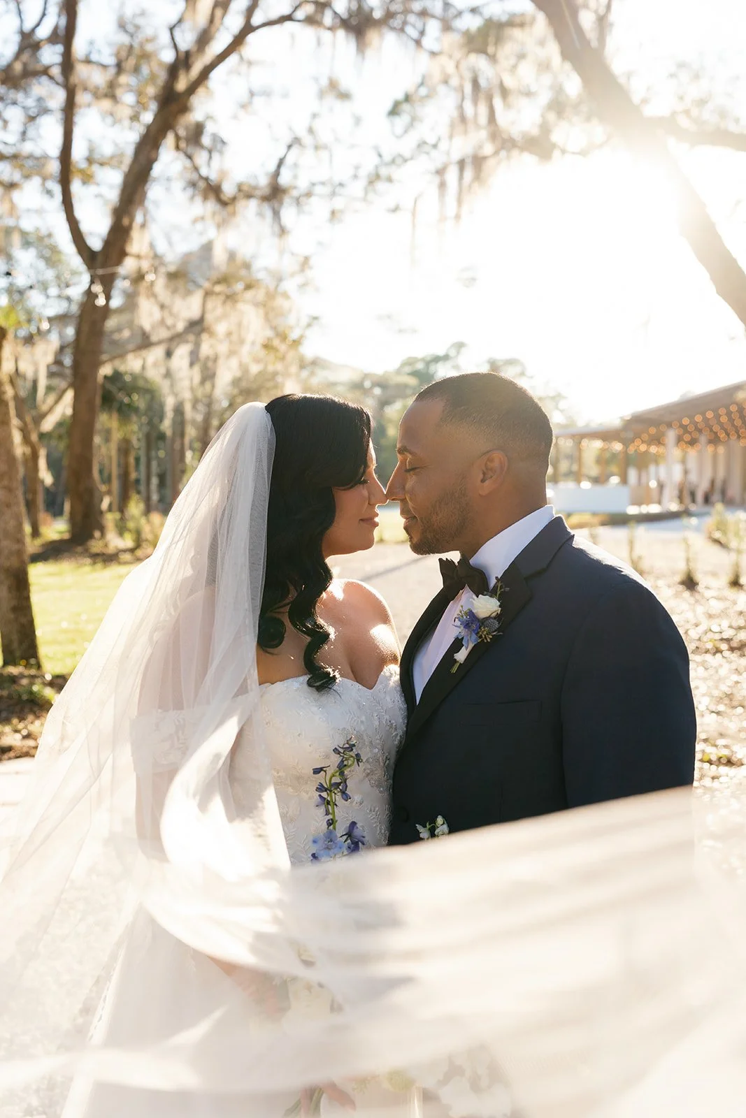 A bride and groom face each other outdoors on a sunny day, dressed in wedding attire.