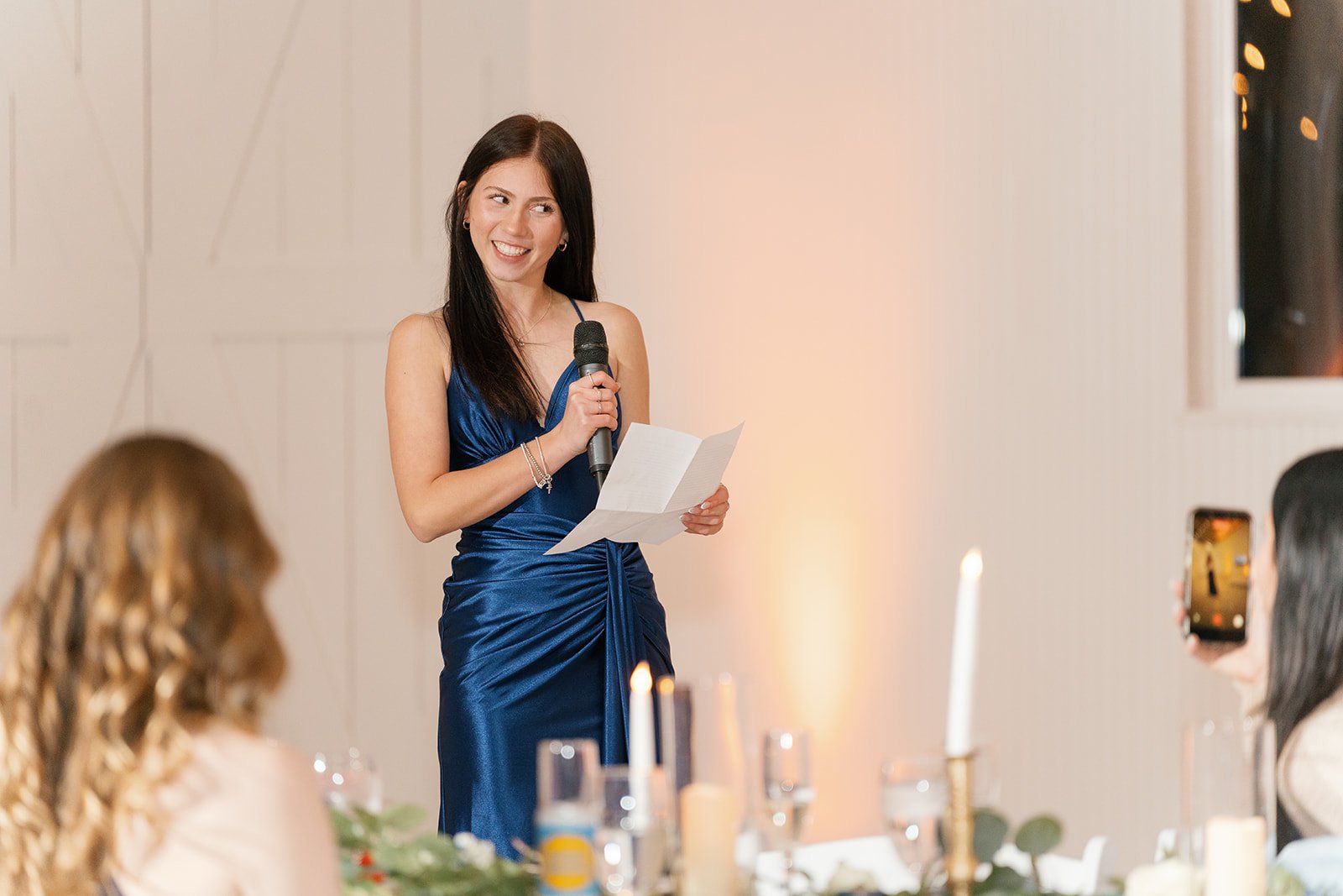 A woman in a shiny blue dress is speaking into a microphone at a formal event, holding a paper, with guests listening and taking pictures in a decorated venue.