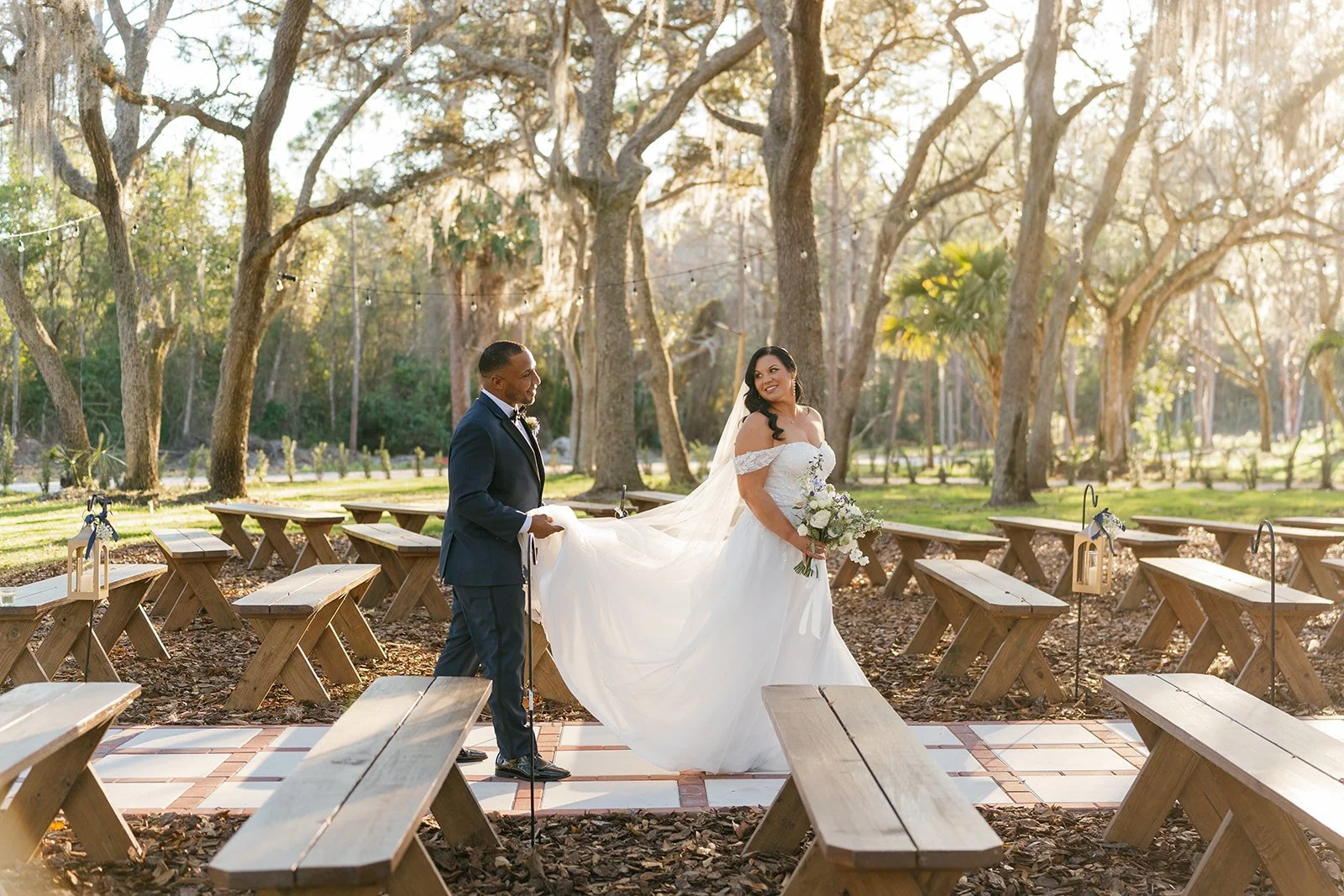 A bride in a white wedding gown holding a bouquet of white flowers, smiling at a groom in a navy suit, in an outdoor setting with trees and sunlight, surrounded by wooden benches.