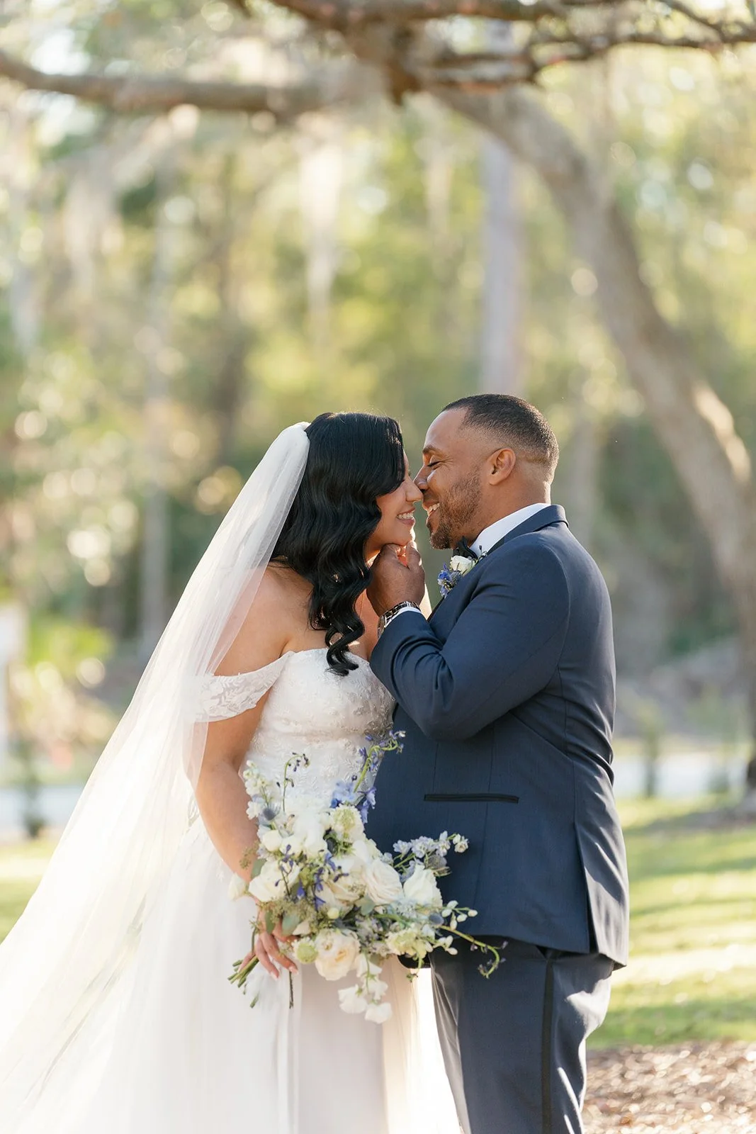 A bride and groom are standing close together outdoors, smiling and touching foreheads, with a blurred natural background of trees. The bride is holding a bouquet of white and purple flowers, and she is wearing an off-the-shoulder wedding dress with 
