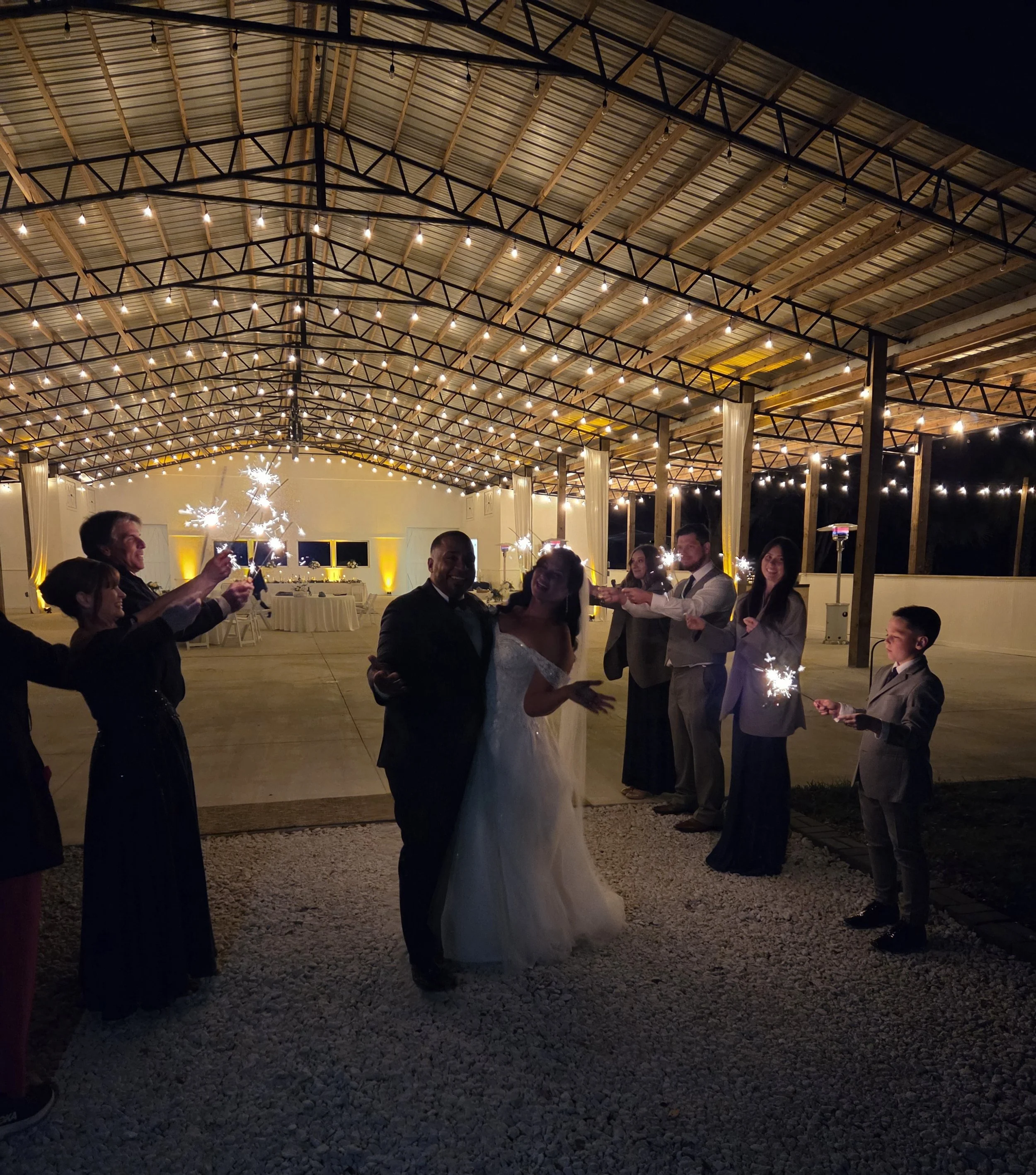 Scene of a wedding celebration at night, with guests holding sparklers under a structure with string lights and a high metal roof, and a bride and groom dancing.