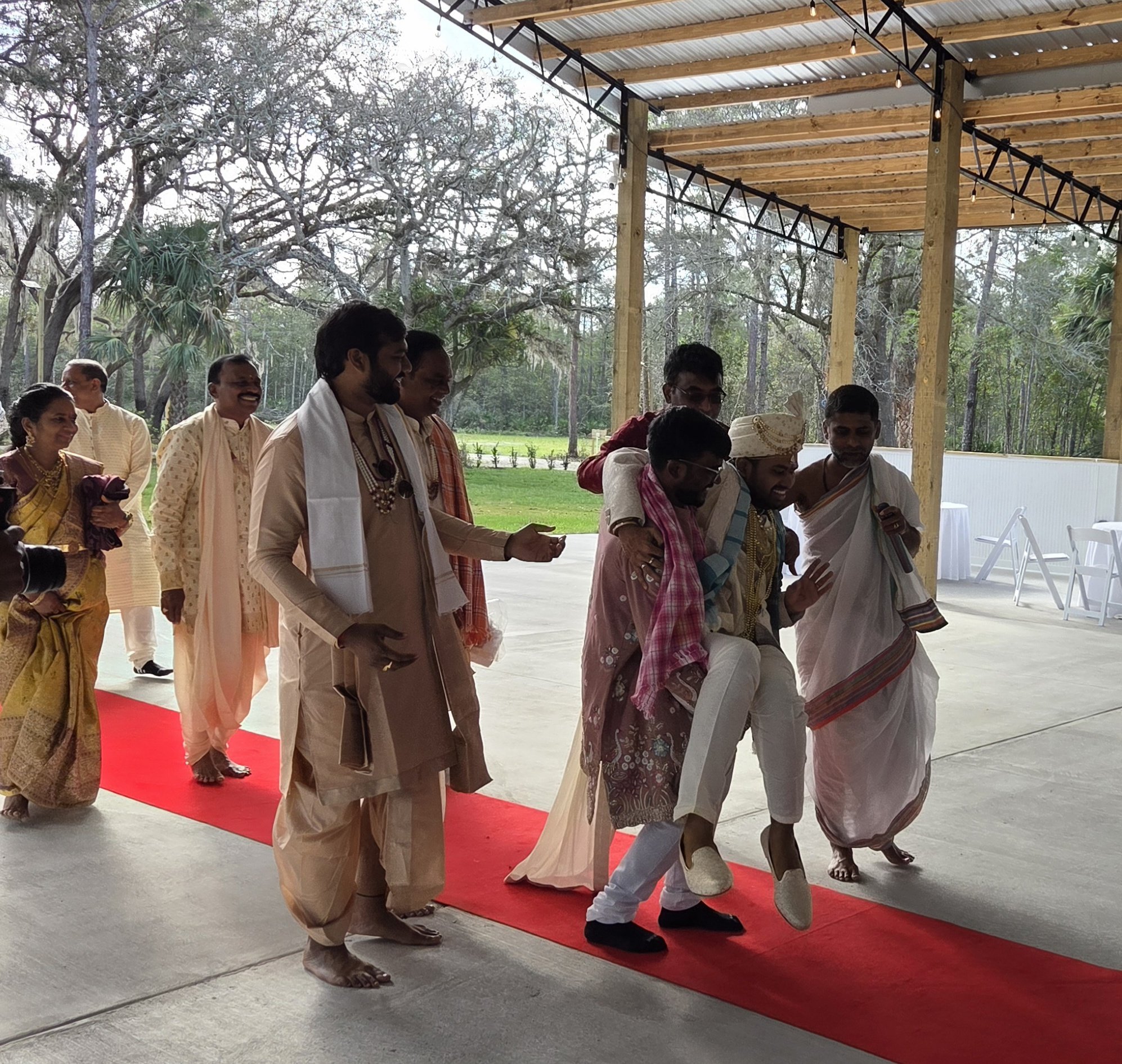 Group of people, dressed in traditional Indian clothing, standing on a red carpet under a wooden and metal canopy, engaged in a prayer or ceremony outdoors during daytime.