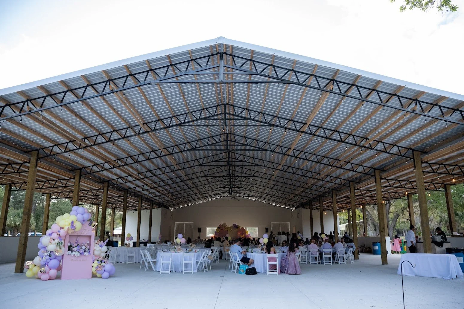 Indoor event space with a high metal roof, decorated tables with pink and white balloons, and guests seated at round tables for a celebration or party.