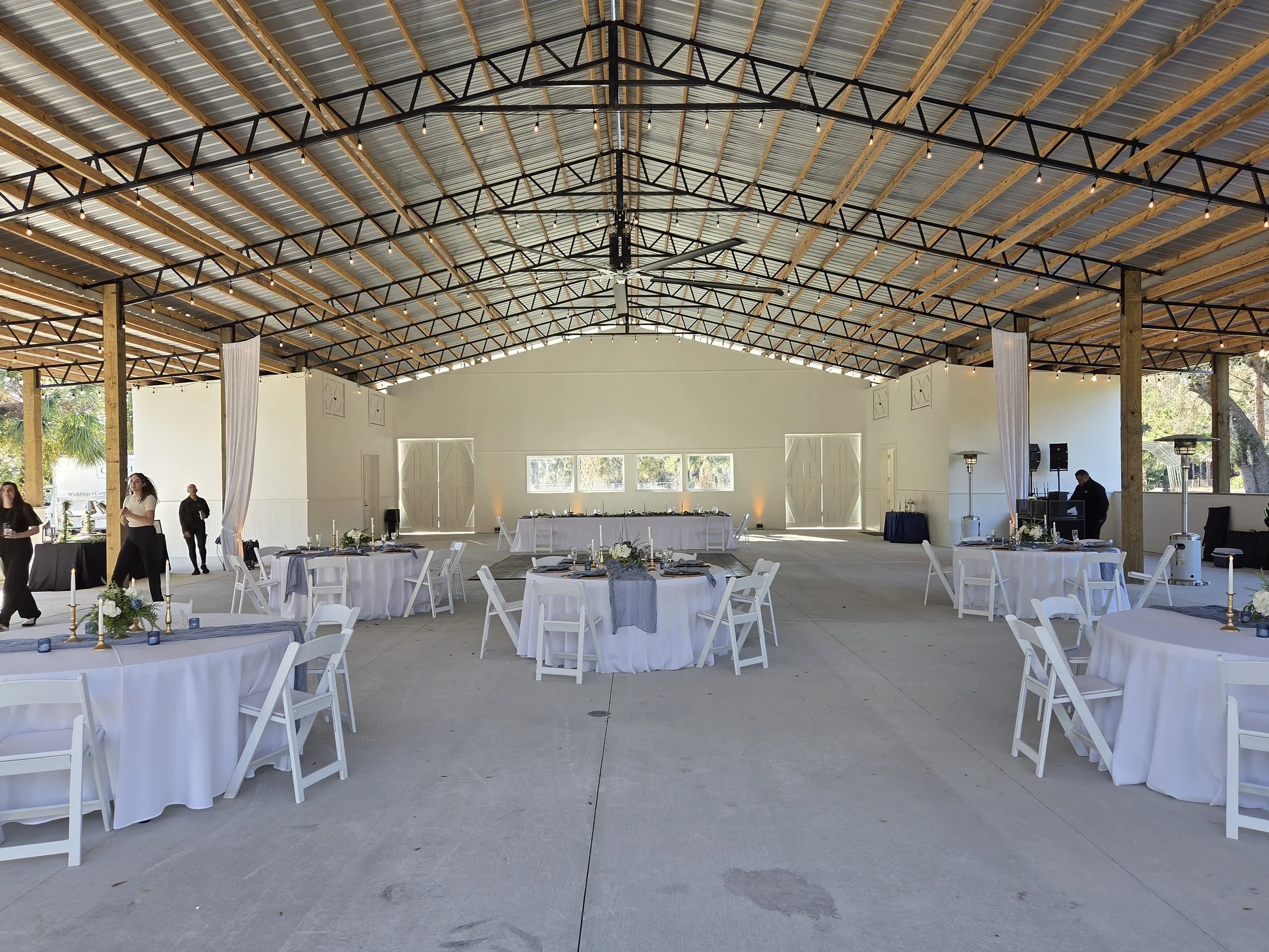Indoor event space decorated with white tables and chairs, floral centerpieces, and candles, with a high metal and wood ceiling, and large windows on the back wall, set for a celebration or reception.