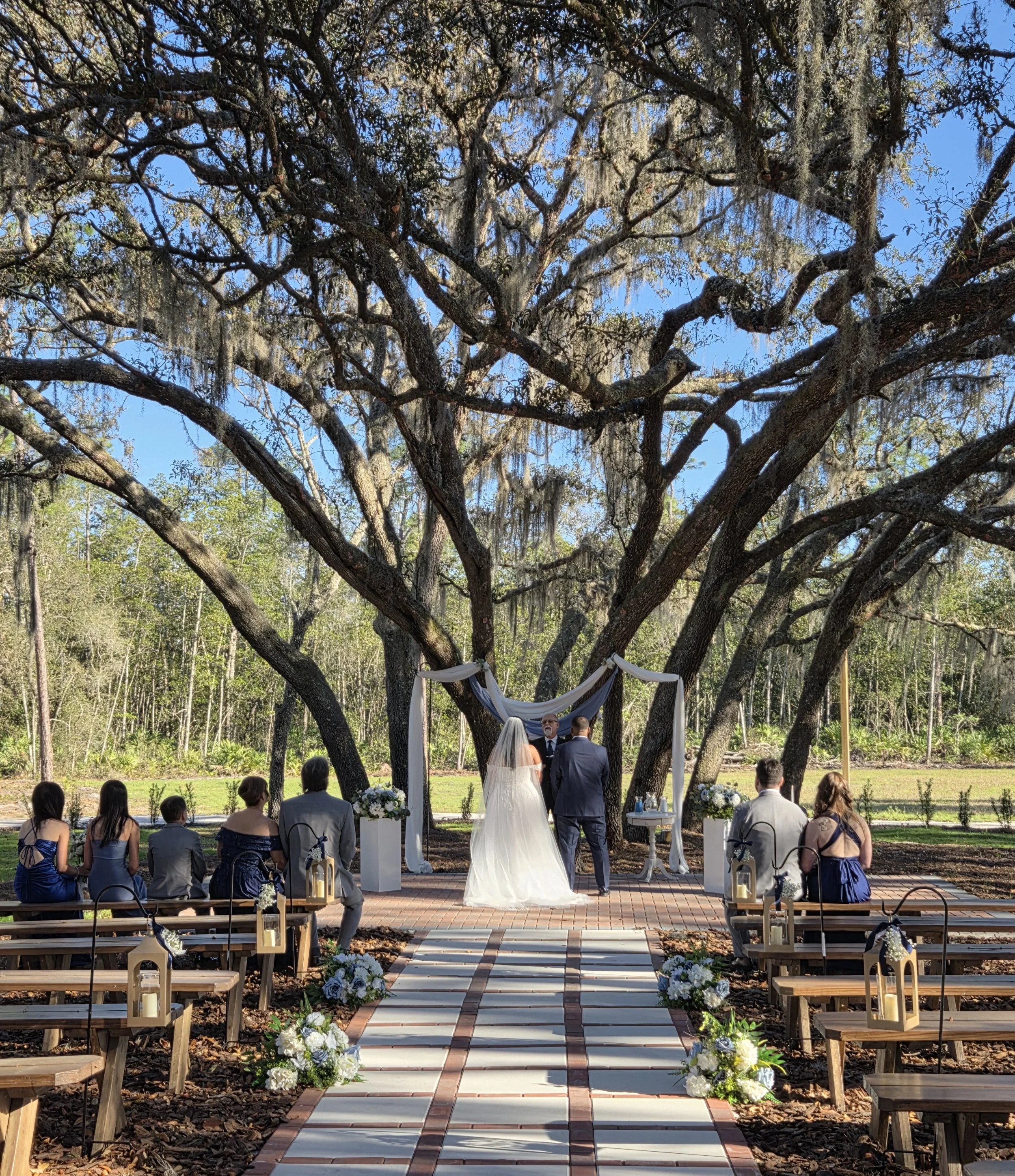 A wedding ceremony taking place outdoors beneath a large tree with hanging moss. A bride in a white gown and veil stands next to the groom in a suit, with an officiant in front of them. Guests are seated on wooden benches decorated with flowers, faci