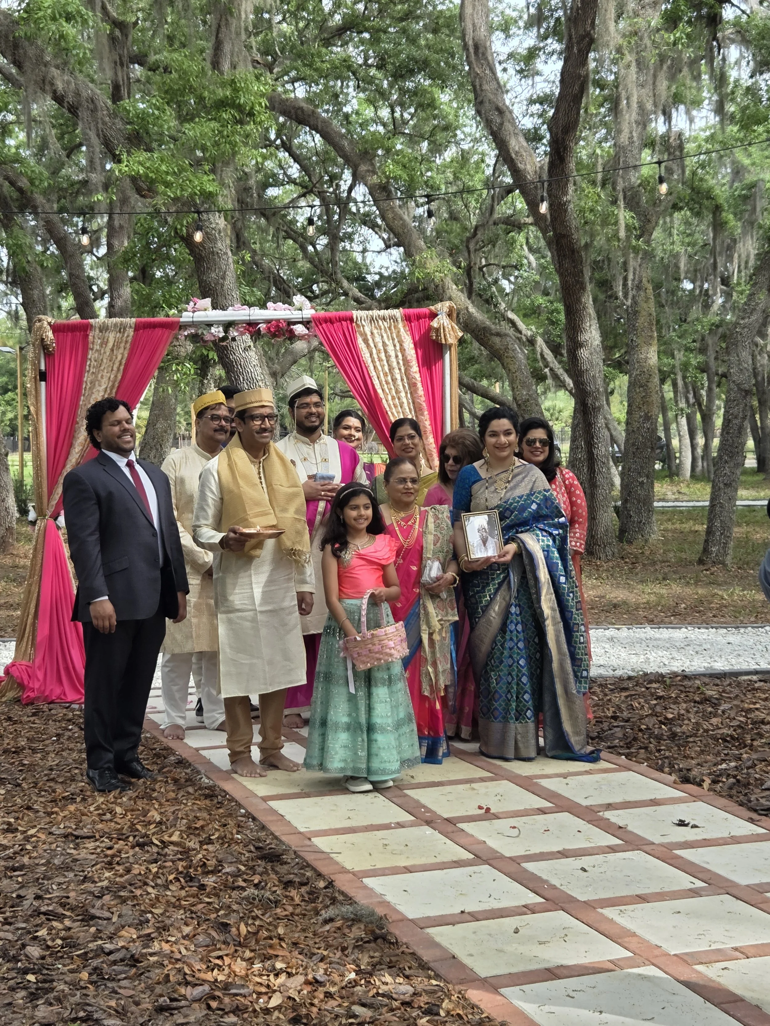 A group of people dressed in traditional Indian attire at an outdoor gathering, standing on a checkered pathway under trees, with pink and gold drapery and string lights in the background.