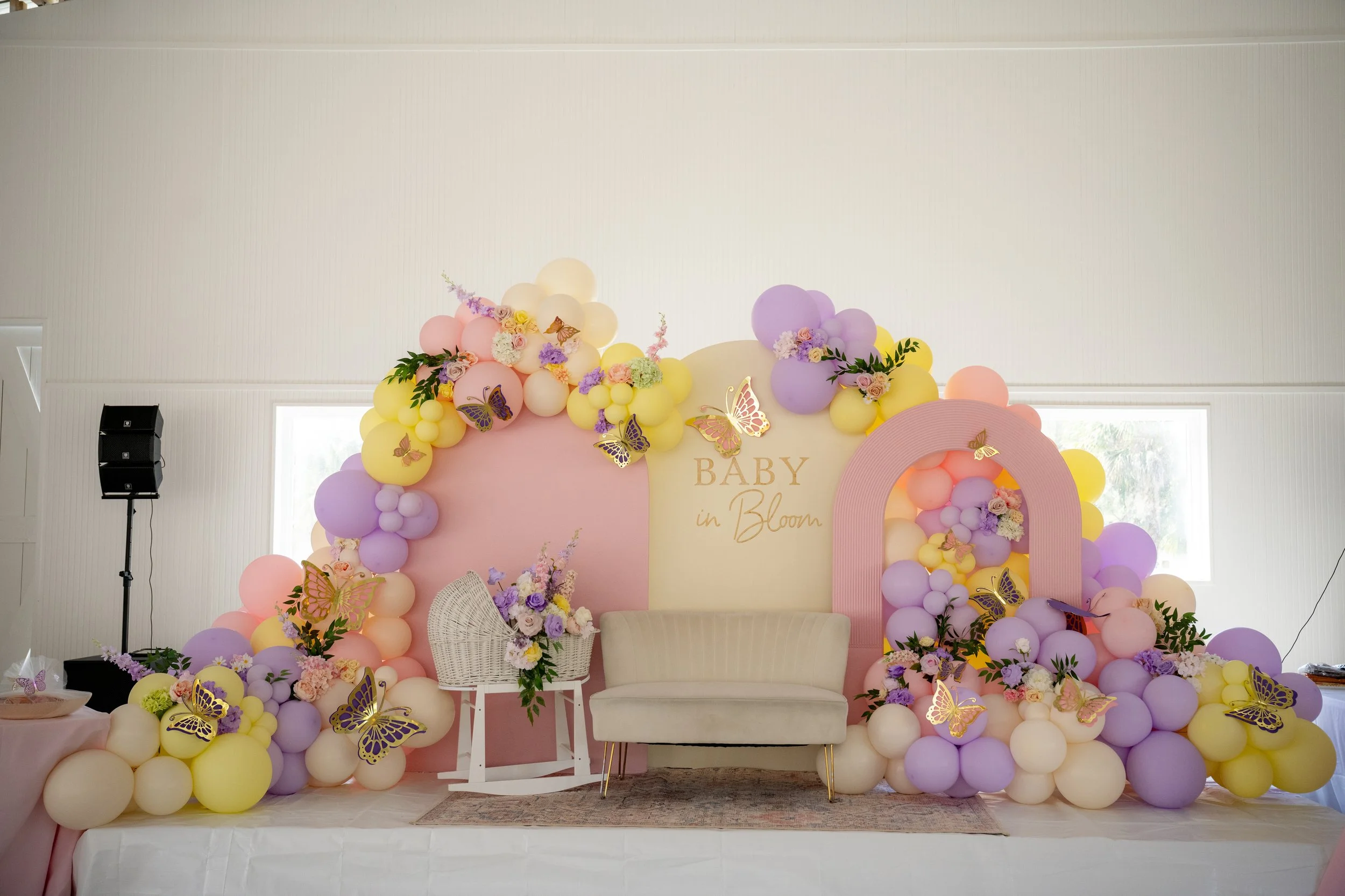 Decorative backdrop for a baby shower with pastel pink, yellow, and purple balloons, floral arrangements, and butterfly decorations. The backdrop features the phrase 'Baby in Bloom' and includes a white wicker chair and a white cushioned chair in fro