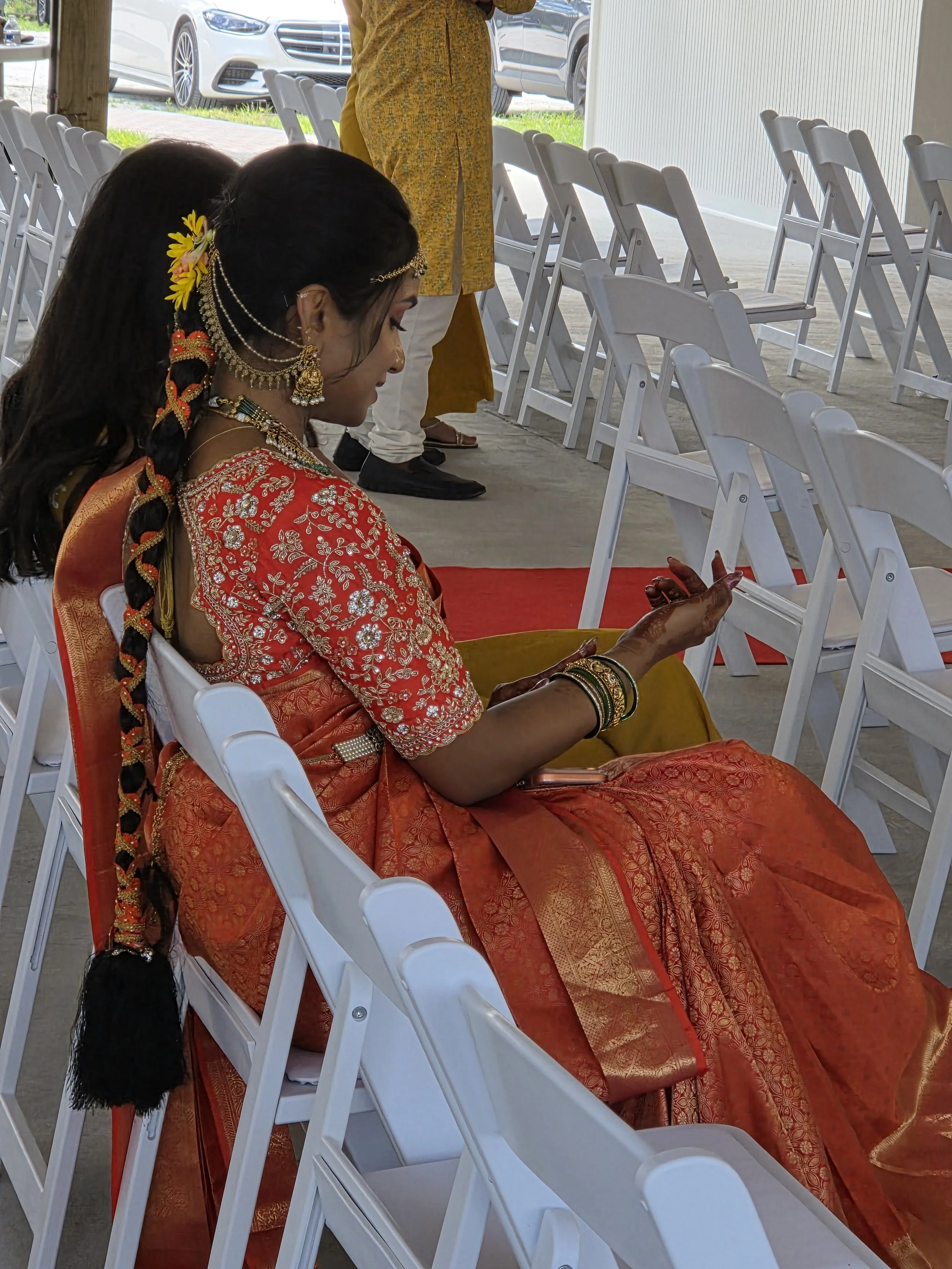 A woman dressed in traditional Indian attire, including a red and gold embroidered saree and intricate jewelry, sitting on a white chair at an event, with a row of empty chairs nearby and cars visible outside.