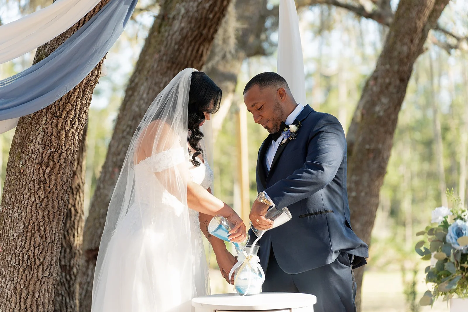 A bride and groom performing a sand pouring ceremony during an outdoor wedding in the woods, with trees and greenery in the background.