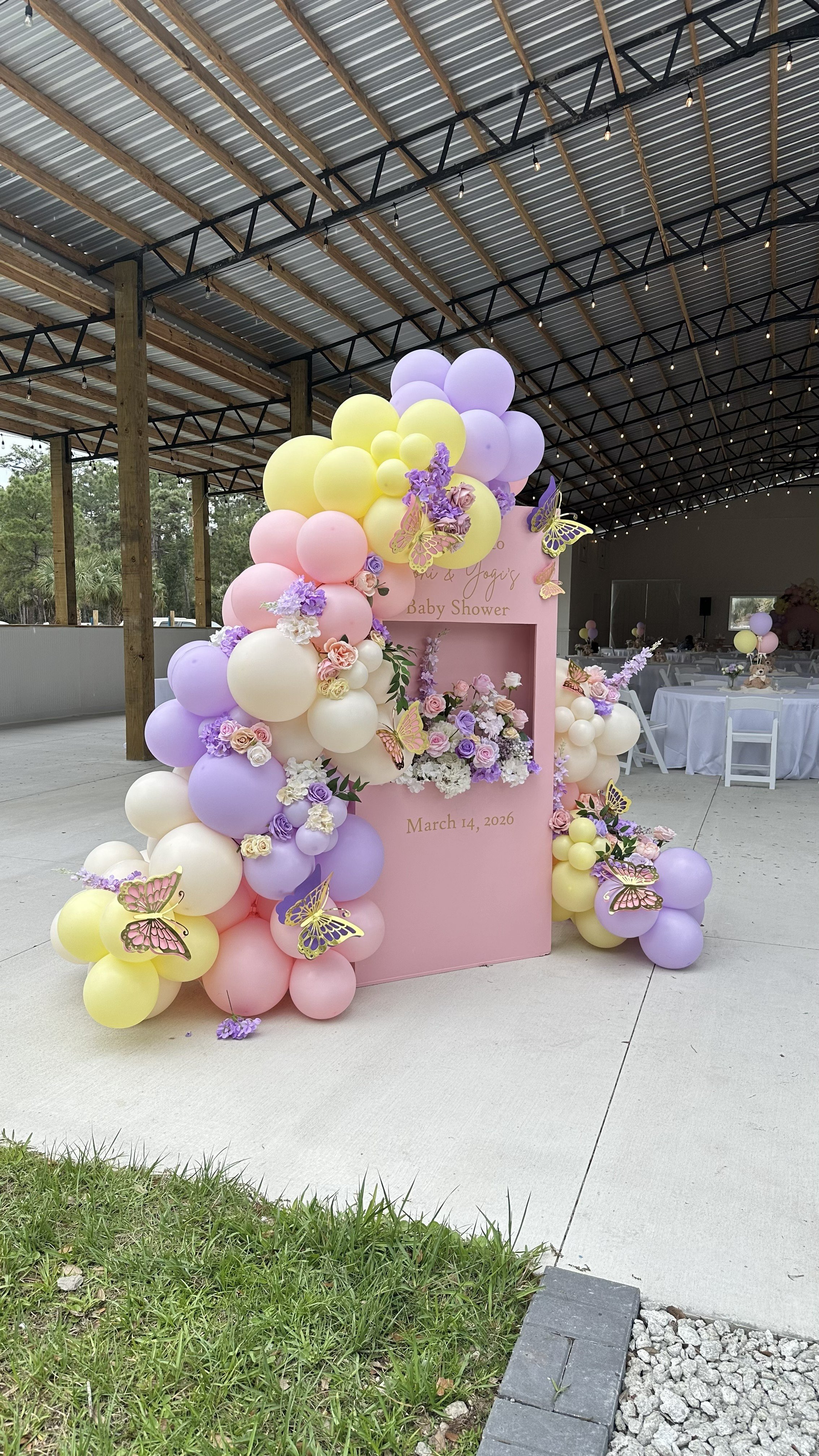 Decorative pink baby shower arch with pastel balloons, flowers, and butterfly accents, with a backdrop reading "Baby Shower" and the date March 14, 2026, inside a large open venue with string lights.