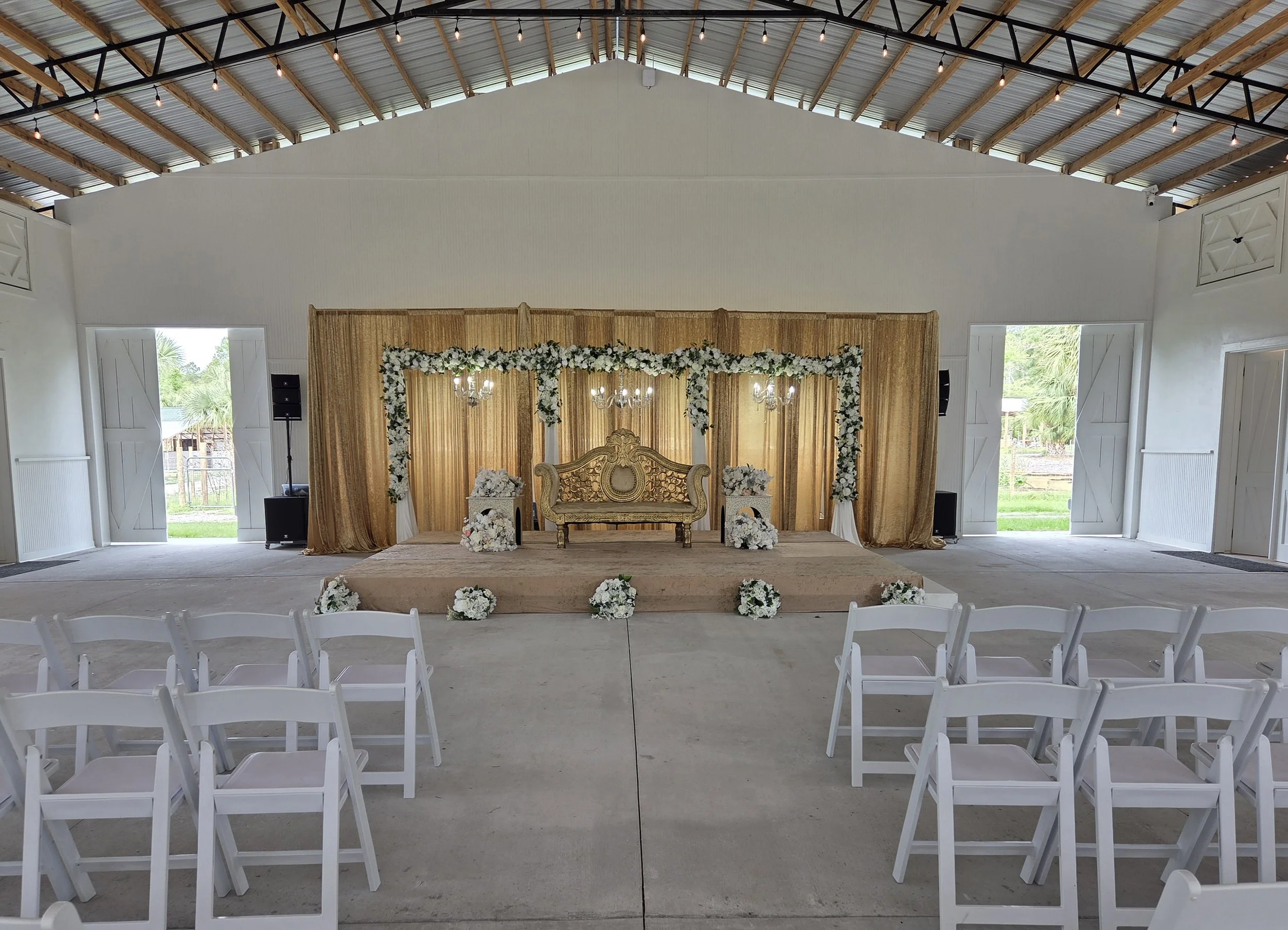 Wedding ceremony setup with a decorated altar, floral arrangements, and rows of chairs inside a rustic barn-style venue with open doors and string lights.
